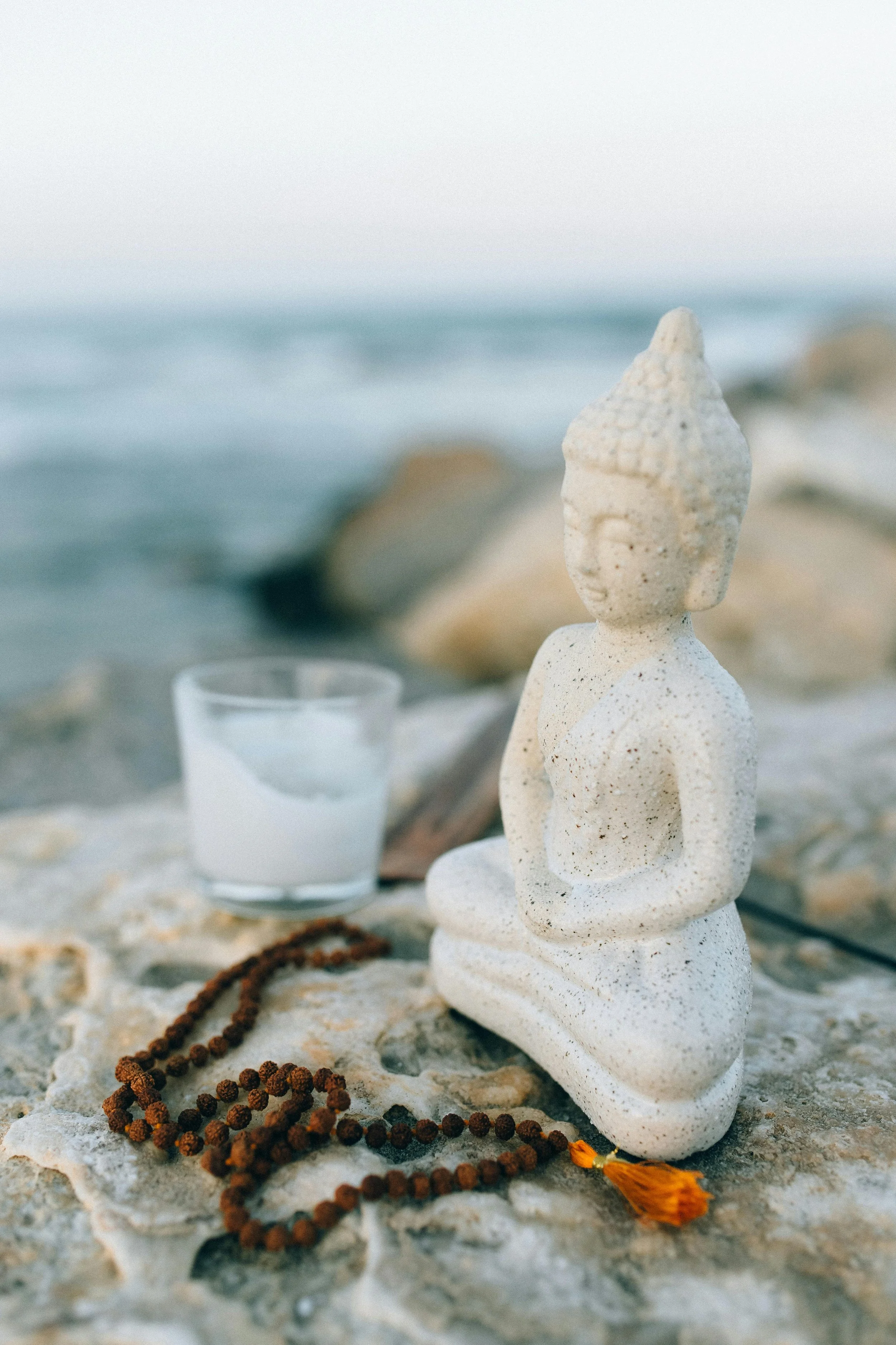 Sand sculpture of a meditating figure with closed eyes on a beach, surrounded by a string of prayer beads, a glass of white liquid, rocks, and a small orange tassel.