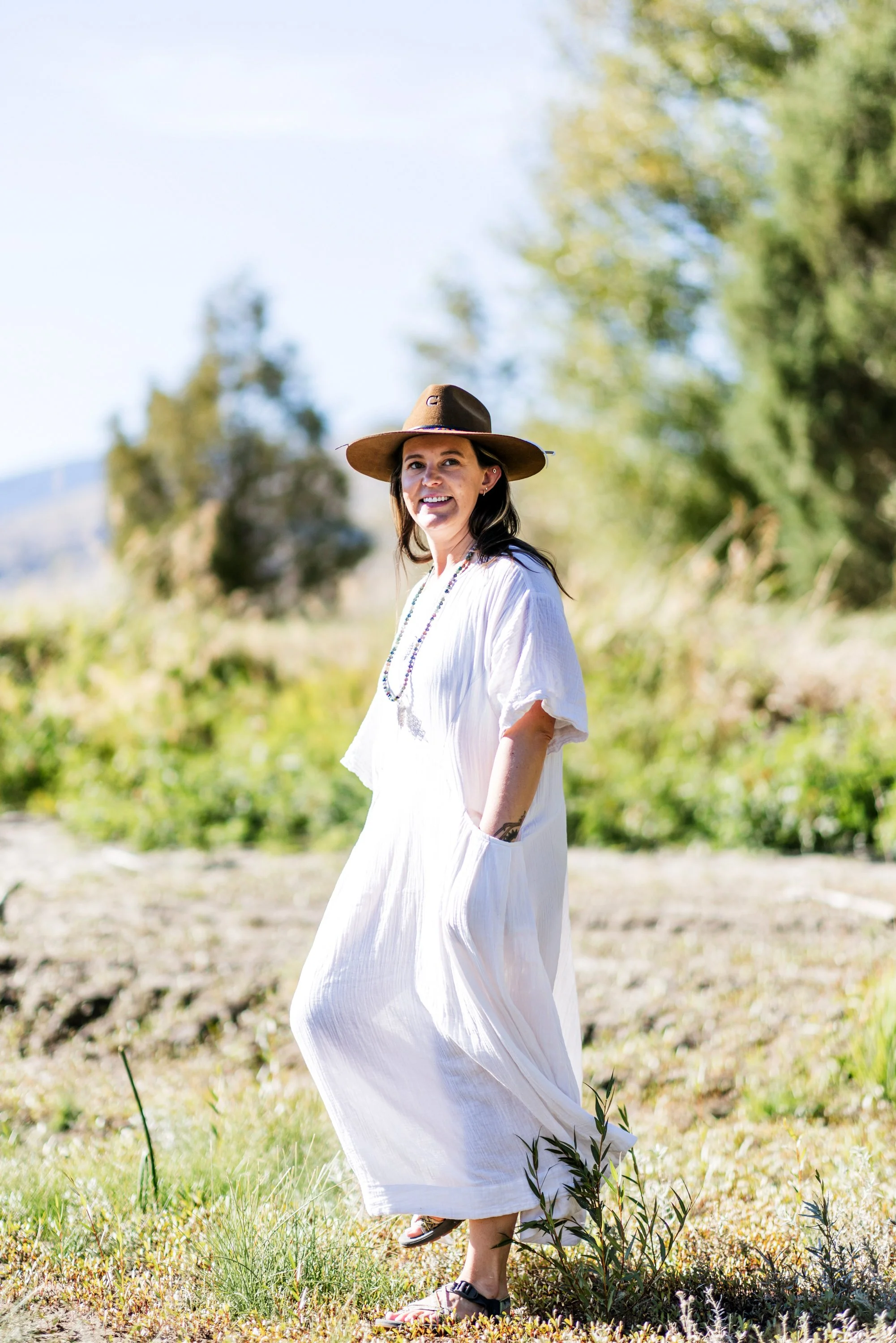 Lou Rae in a white flowing dress and sandals, wearing a wide-brimmed hat, standing outdoors in a grassy field with trees in the background on a sunny day.