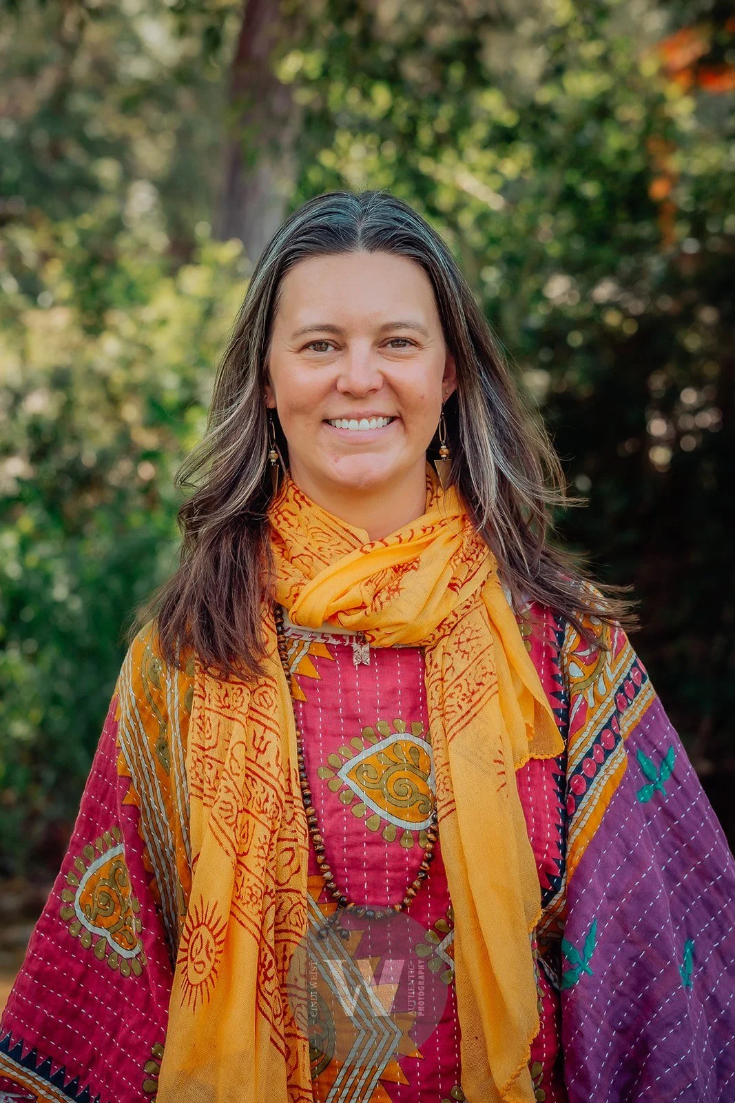 Lou Rae smiling outdoors with a background of green trees, wearing a colorful patterned top and an orange scarf.