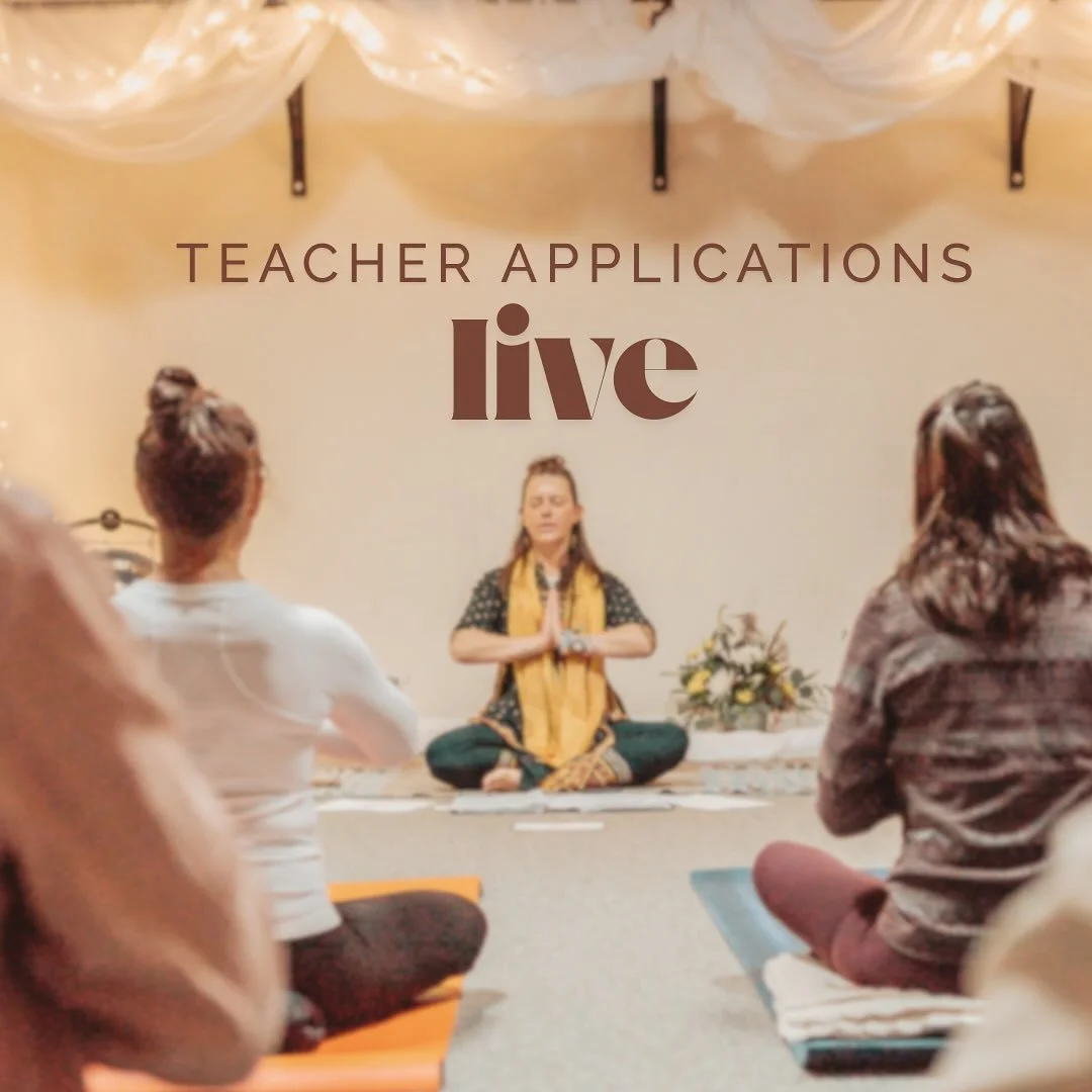 Lou Rae practicing yoga or meditation in a seated, cross-legged position with hands in prayer position, leading a yoga class or workshop in front of a screen that reads 'Teacher Applications Live'.