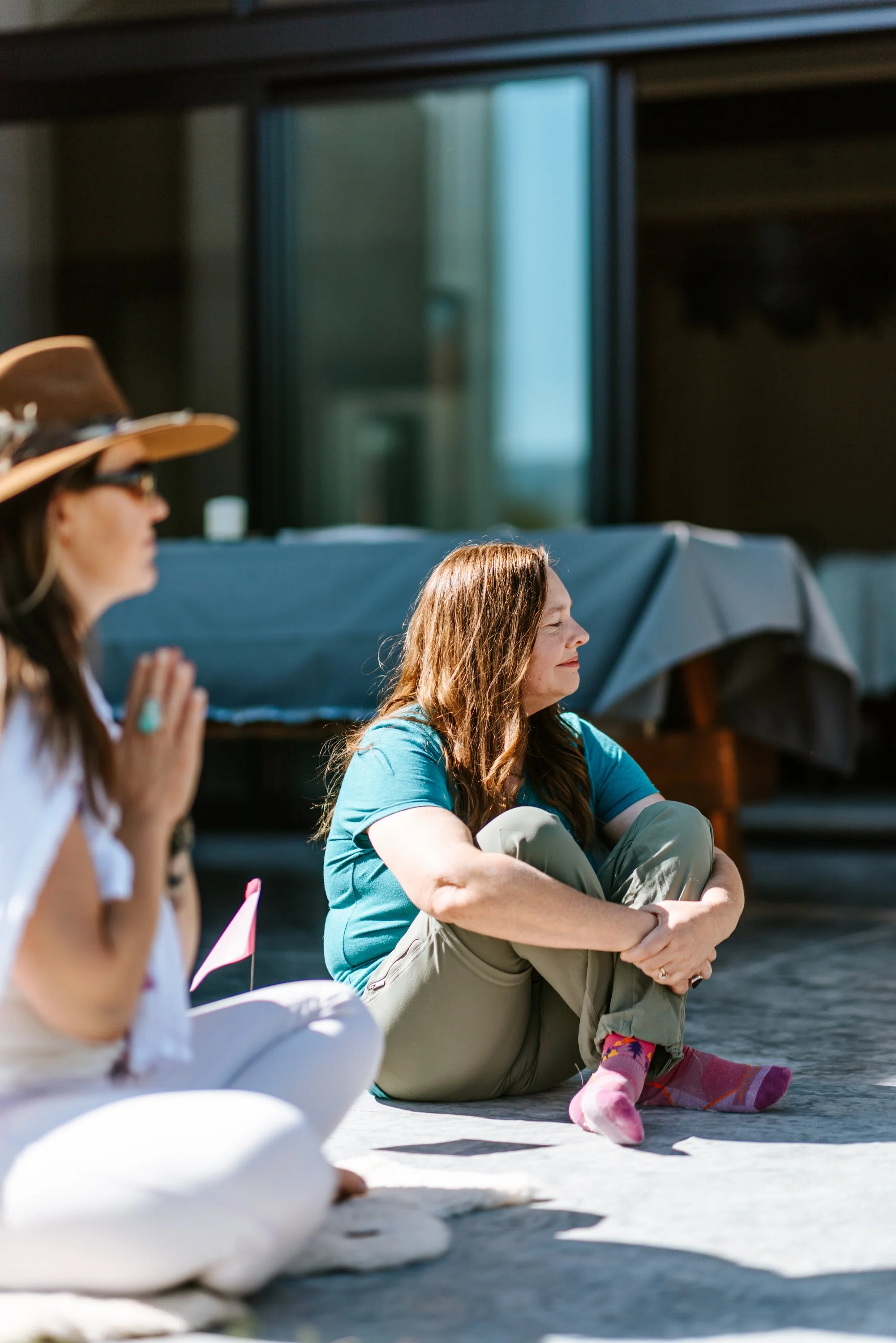 Lou Rae and another woman sitting cross-legged outdoors in sunlight, practicing meditation or mindfulness, with a modern building and umbrellas in the background.