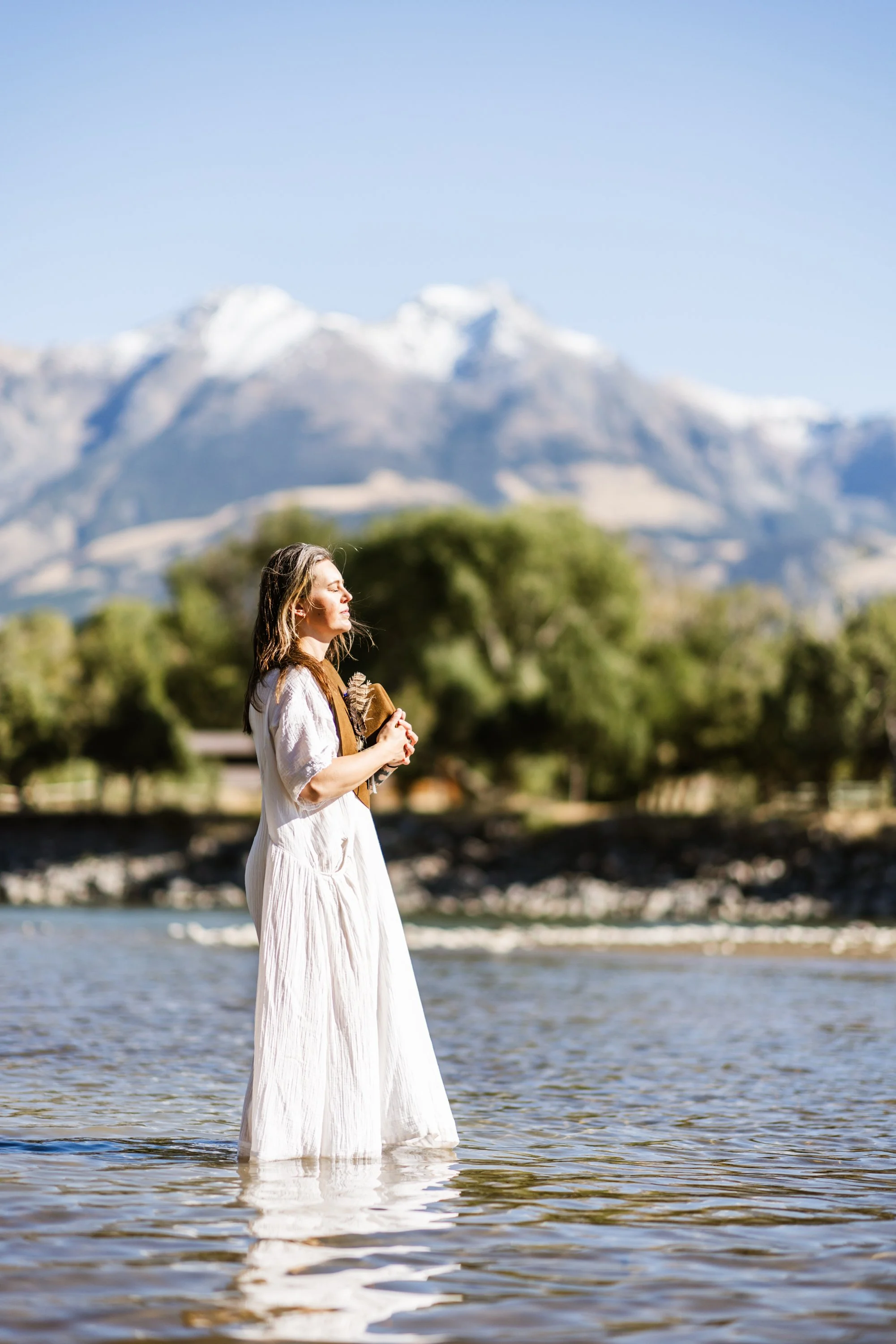 Lou Rae in a white dress standing in shallow water with a mountain range in the background.