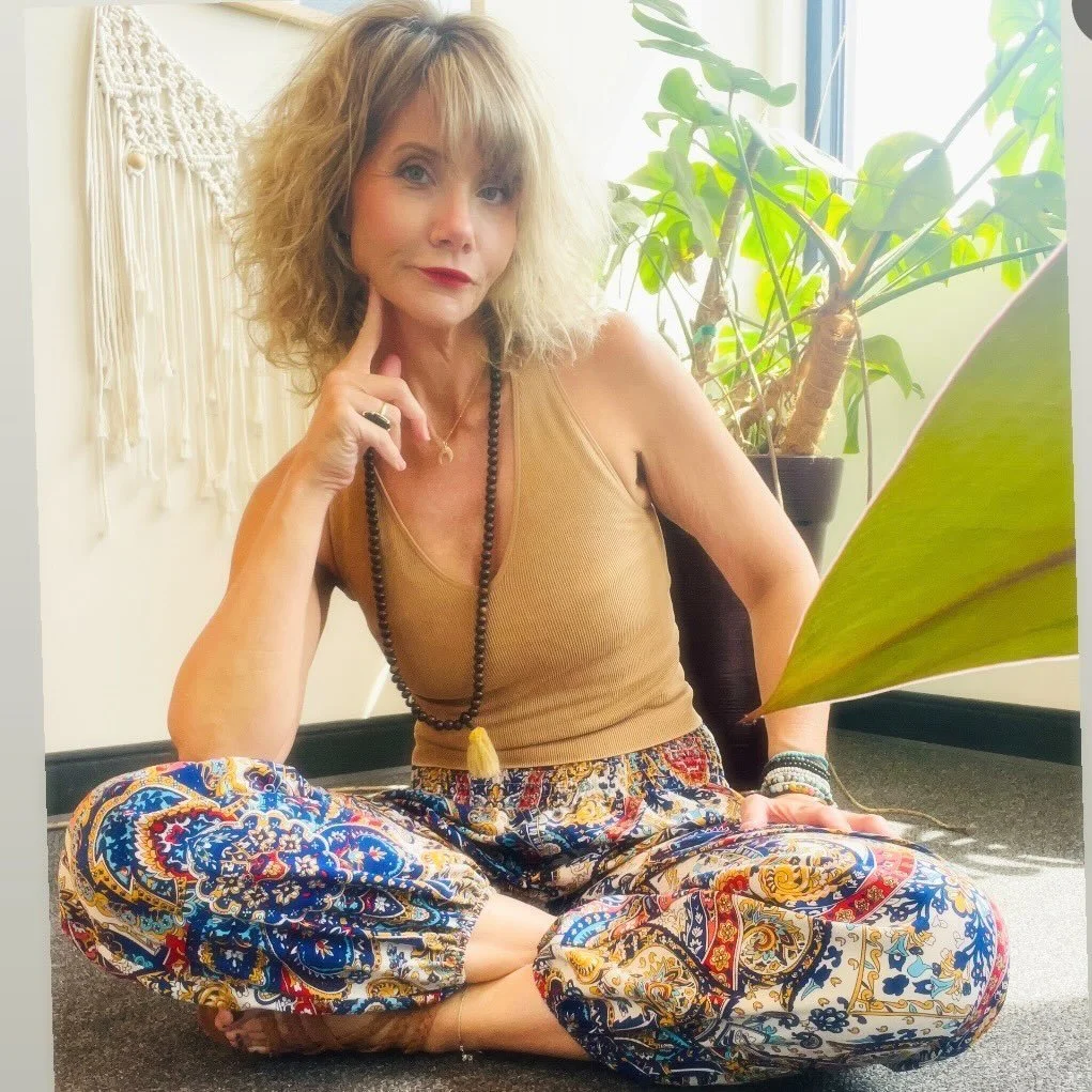A woman with curly blonde hair sitting cross-legged on the floor in a room with natural light, wearing a tan tank top and colorful patterned pants, with one hand resting on her chin and the other on her knee. There are green plants behind her and a wall hanging to her left.