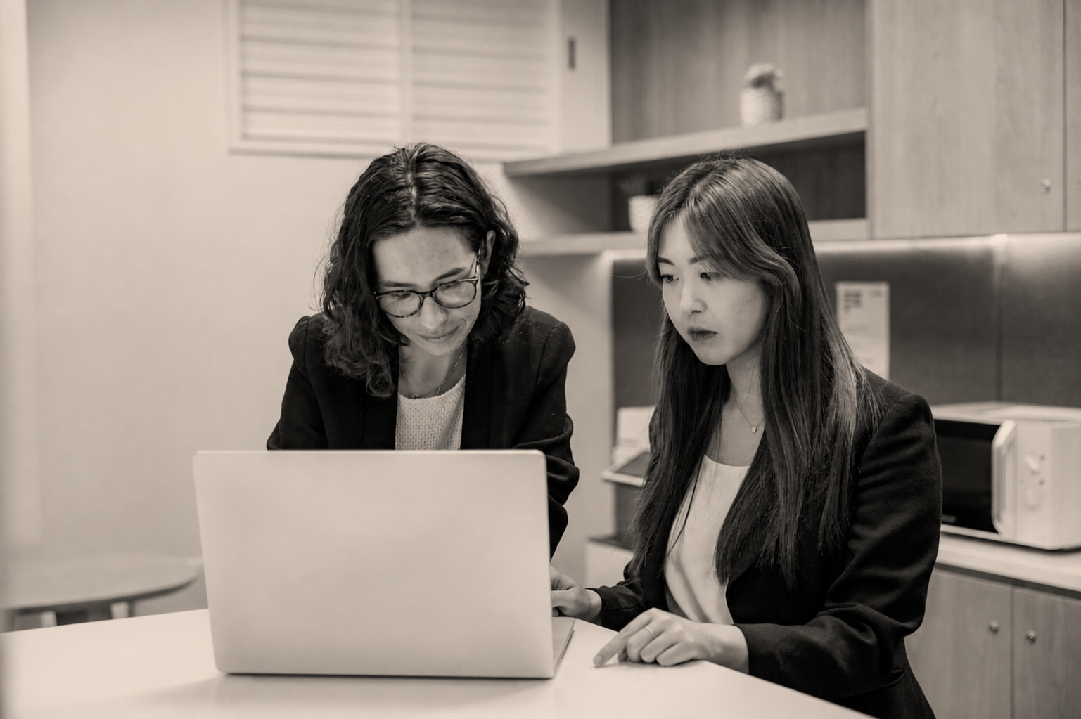 Two women, one with glasses, sitting at a table and looking at a laptop in an office setting.