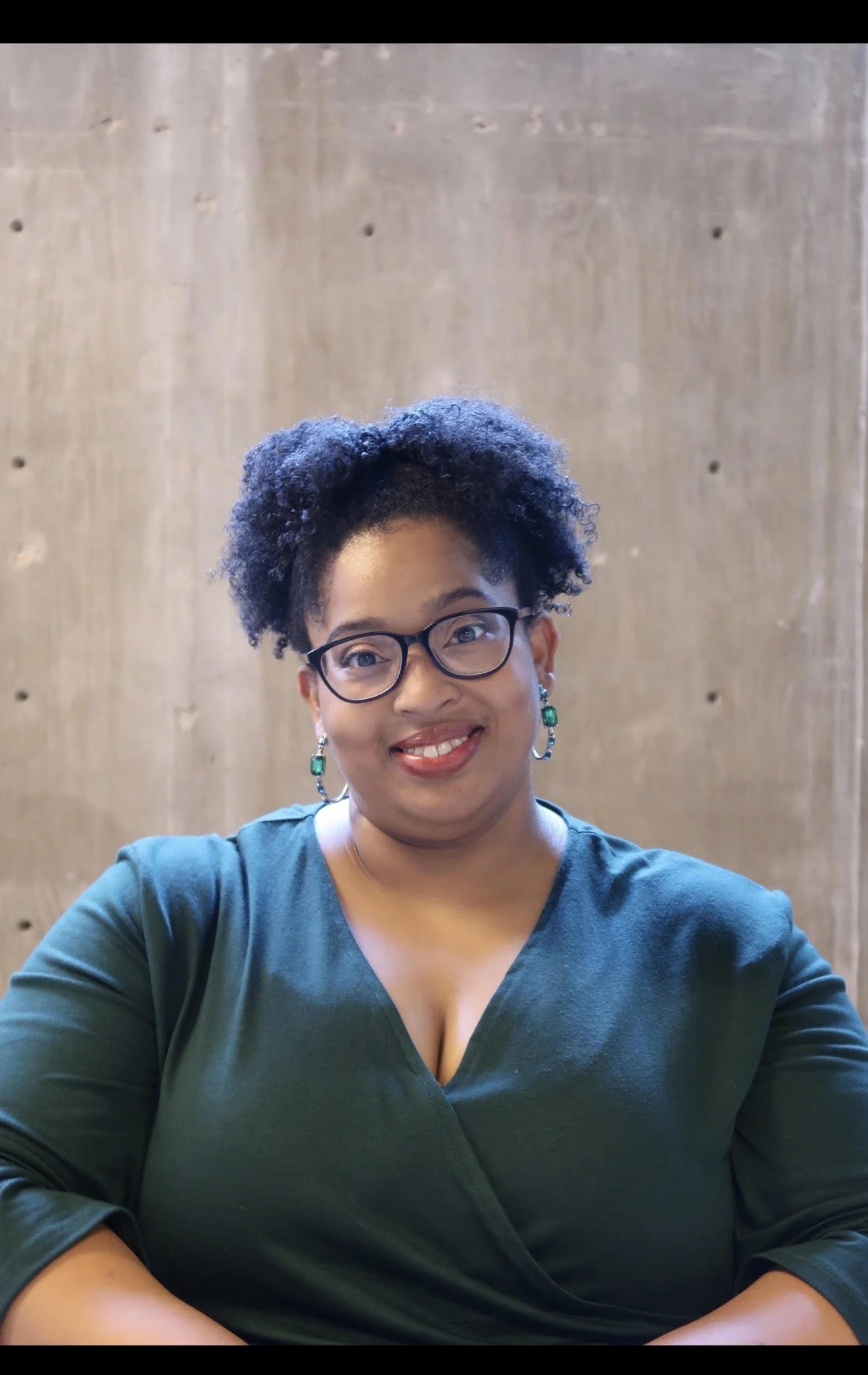 A woman wearing glasses and earrings with green stones, smiling, wearing a dark green top, sitting in front of a wooden wall.