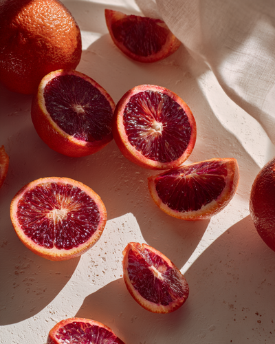 Sliced blood oranges on a white surface with some whole oranges in the background and natural sunlight.