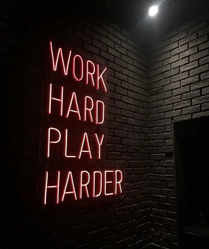 Neon sign reading 'Work Hard Play Harder' on a brick wall in a dark room.