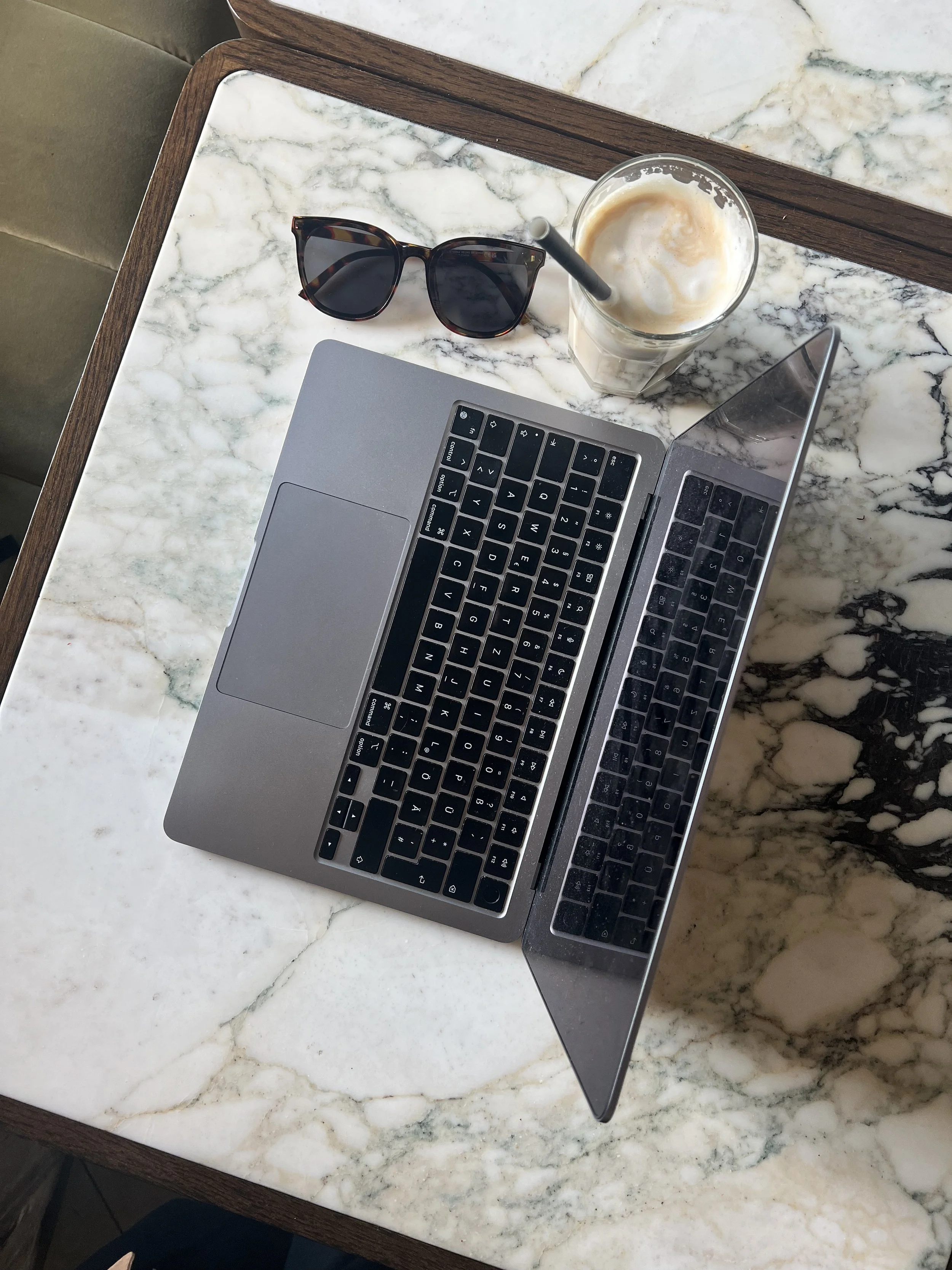 A laptop, a pair of sunglasses, and a iced coffee with a straw on a marble table.