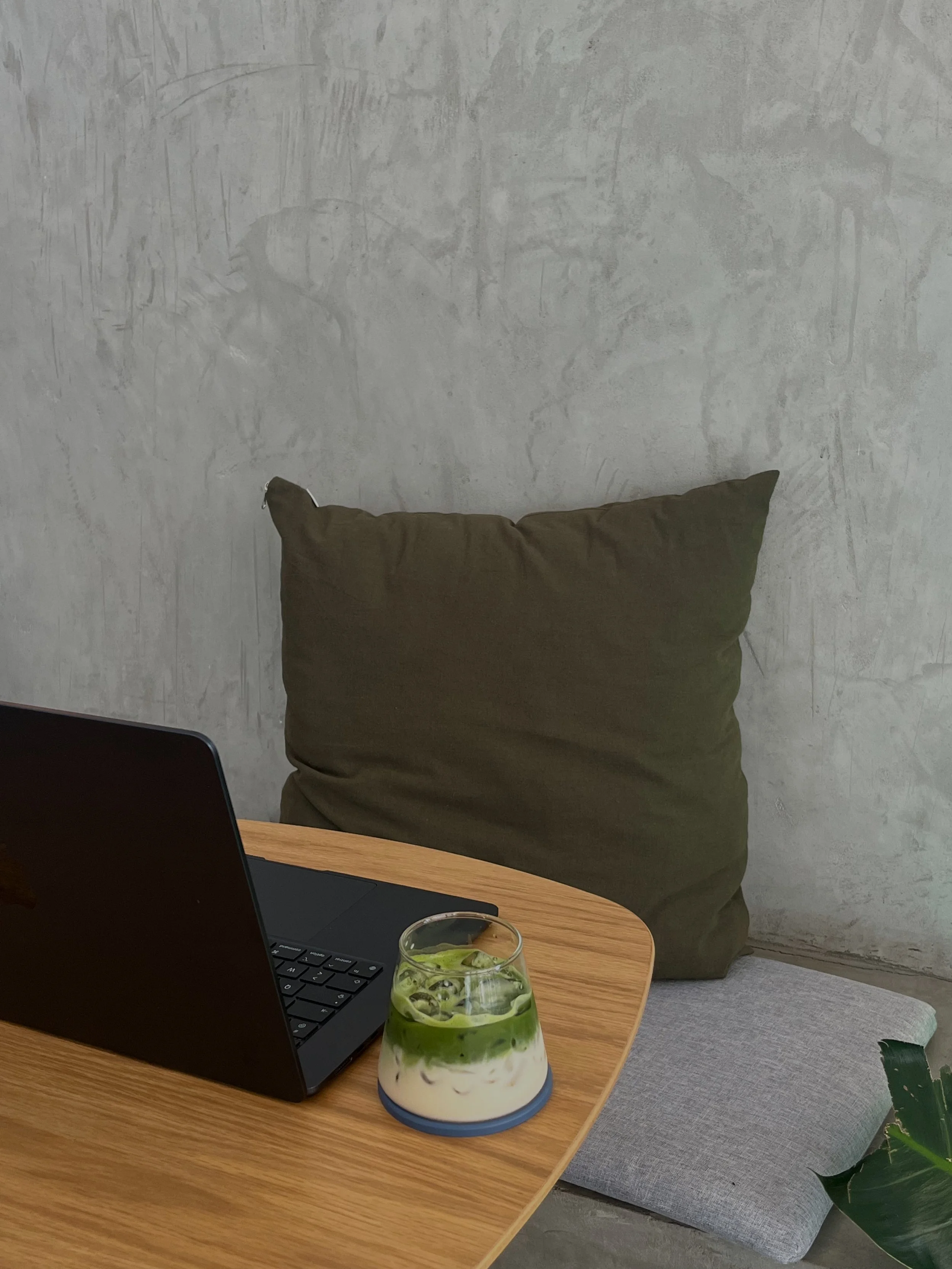 A laptop on a wooden table next to a layered drink with green, white, and brown swirls, a gray cushion, a concrete wall, and a plant in the background.