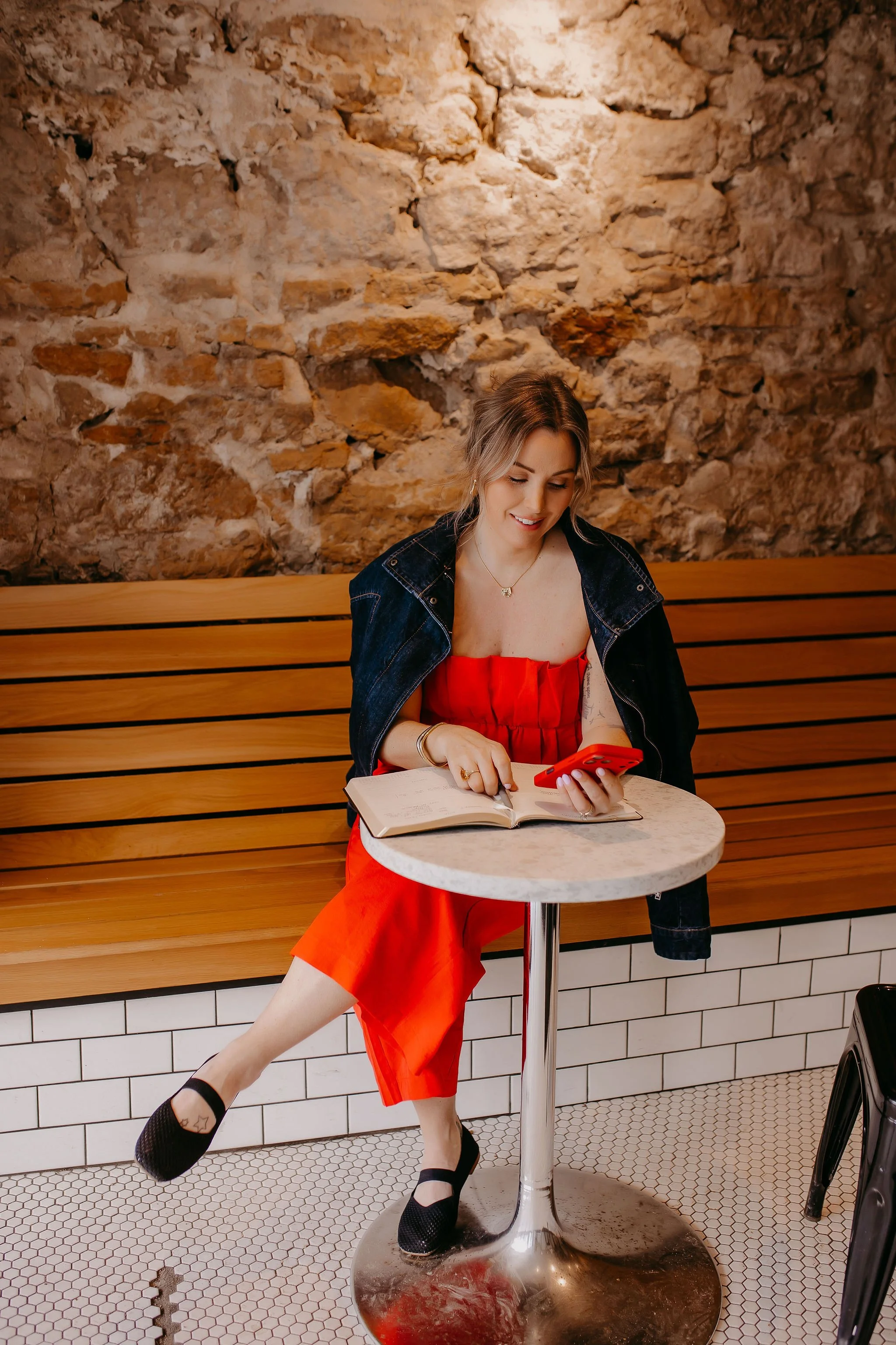A woman sitting on a bench in a cafe, wearing an orange dress, black shoes, with a denim jacket, holding a red phone and a notebook, smiling, with a stone wall in the background.