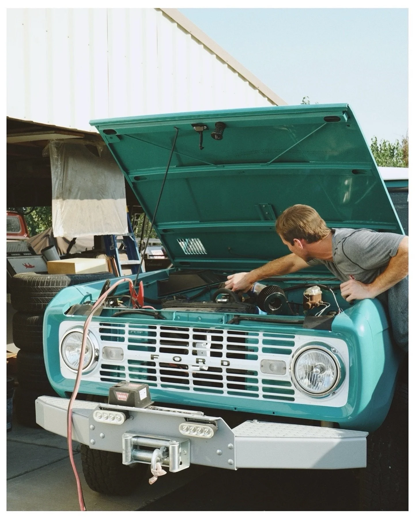 A man working on the engine of a vintage turquoise Ford truck with its hood open, parked in a garage or workshop area.