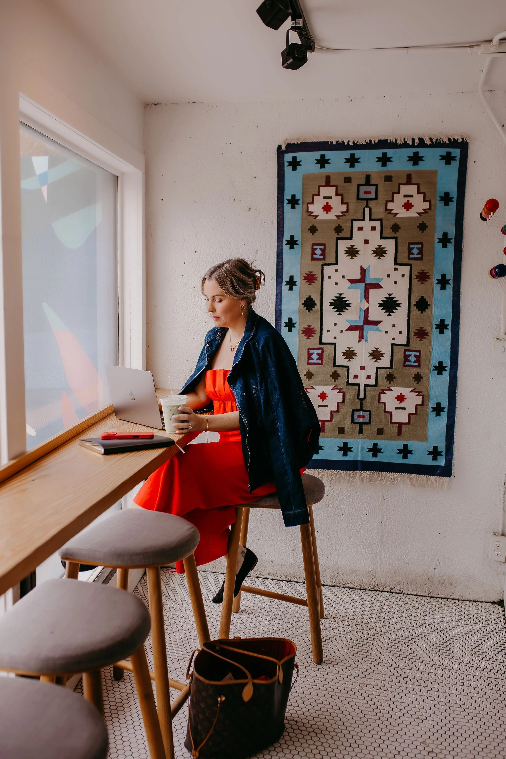 A woman sitting at a wooden counter in a cafe, working on her laptop with a drink in her hand. She is wearing a bright red dress and a black denim jacket. Behind her, a decorative woven rug hangs on the wall, and there are round stools and a patterned tile floor.