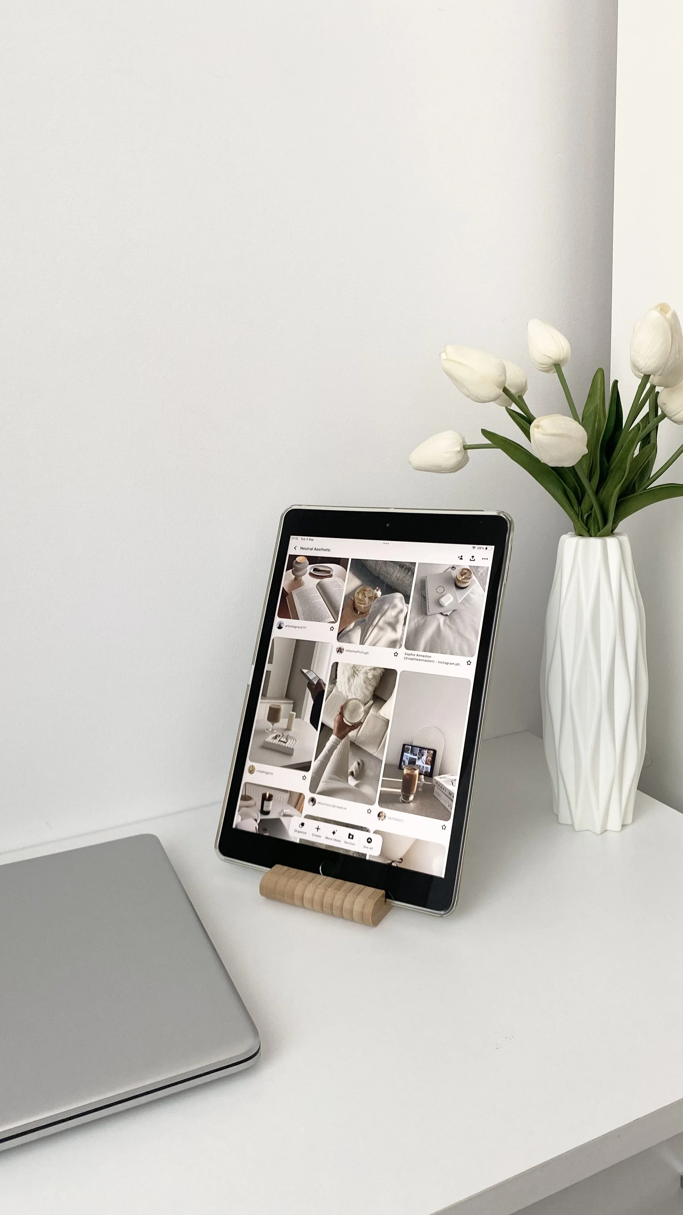 A white desk with a tablet displaying a Pinterest page, a closed silver laptop, and a white vase with white tulips.