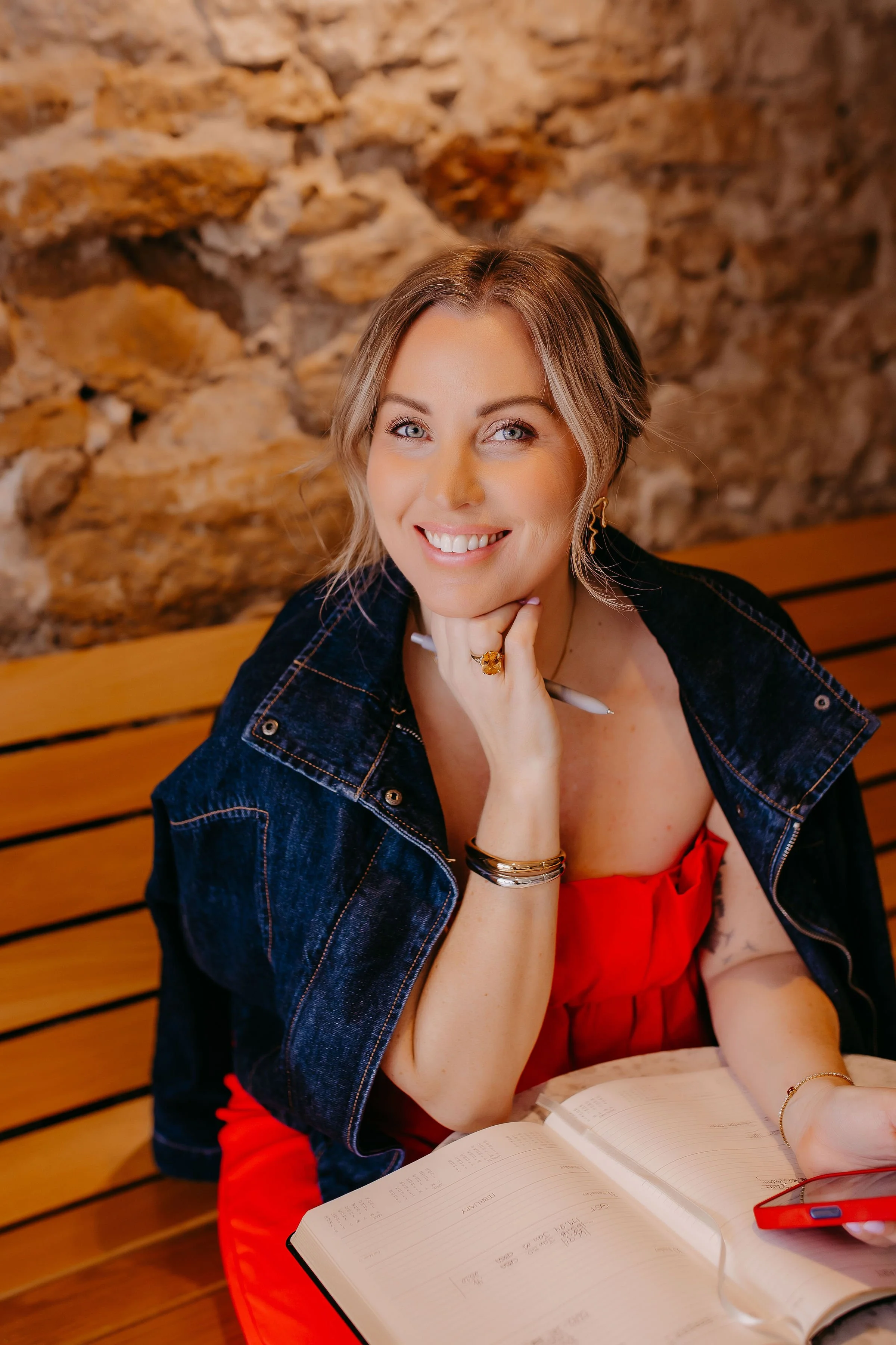 A woman smiling, sitting at a table with an open book, wearing a red dress and denim jacket, with a stone wall background.