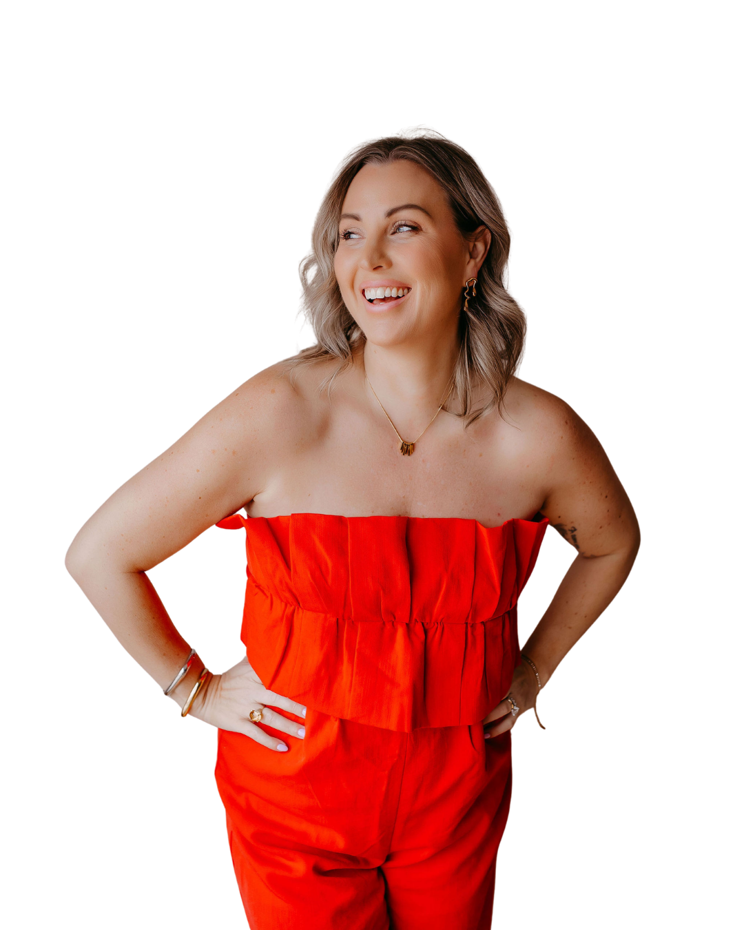 Woman with shoulder-length light brown hair, smiling and looking to her left, wearing a strapless red jumpsuit, gold jewelry, and standing with her hands on her hips.