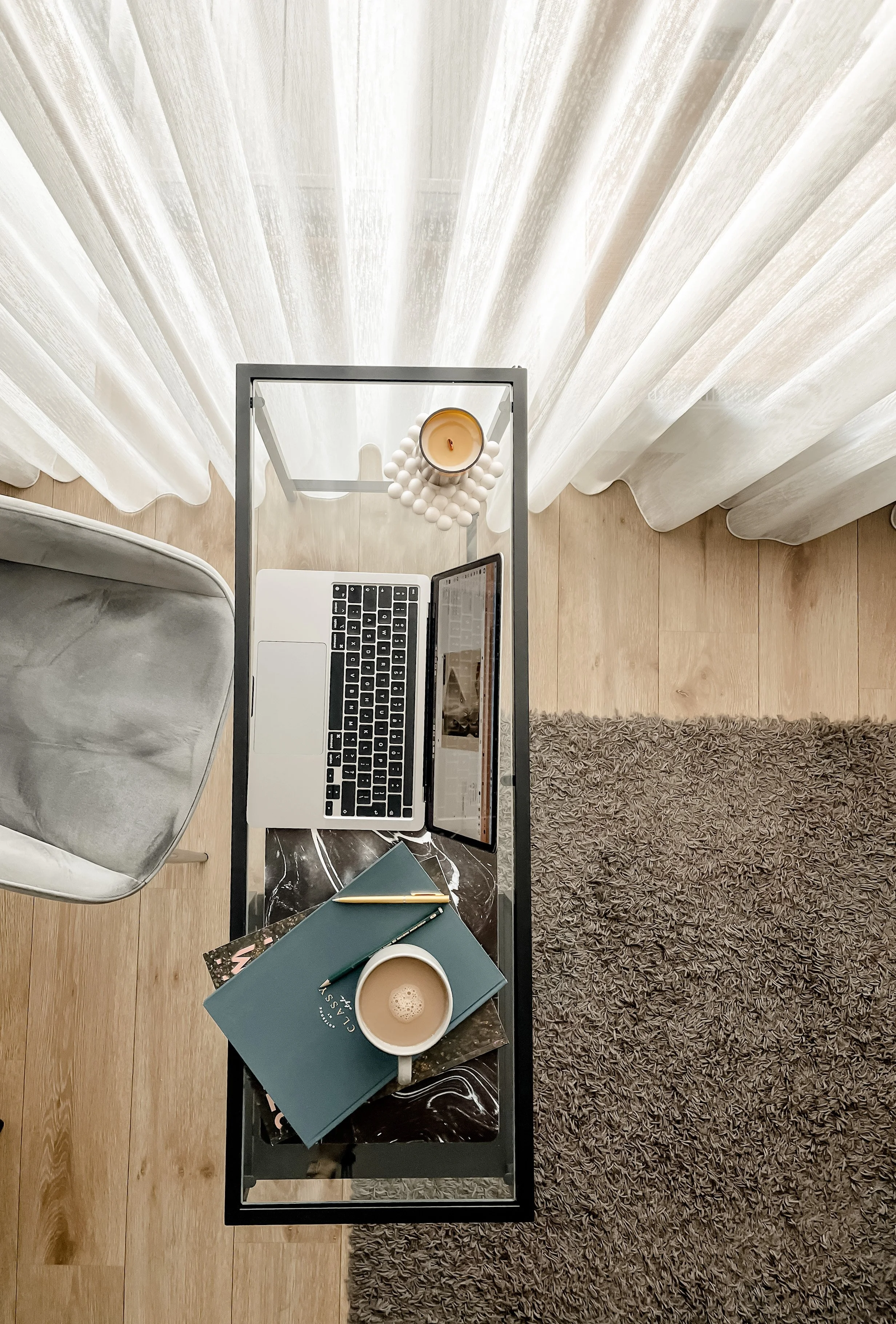 A workspace with a desk that has a laptop, a candle, notebooks, a cup of coffee, and some papers. The desk is positioned in front of sheer curtains, with a chair on the left and a shaggy rug on the floor.