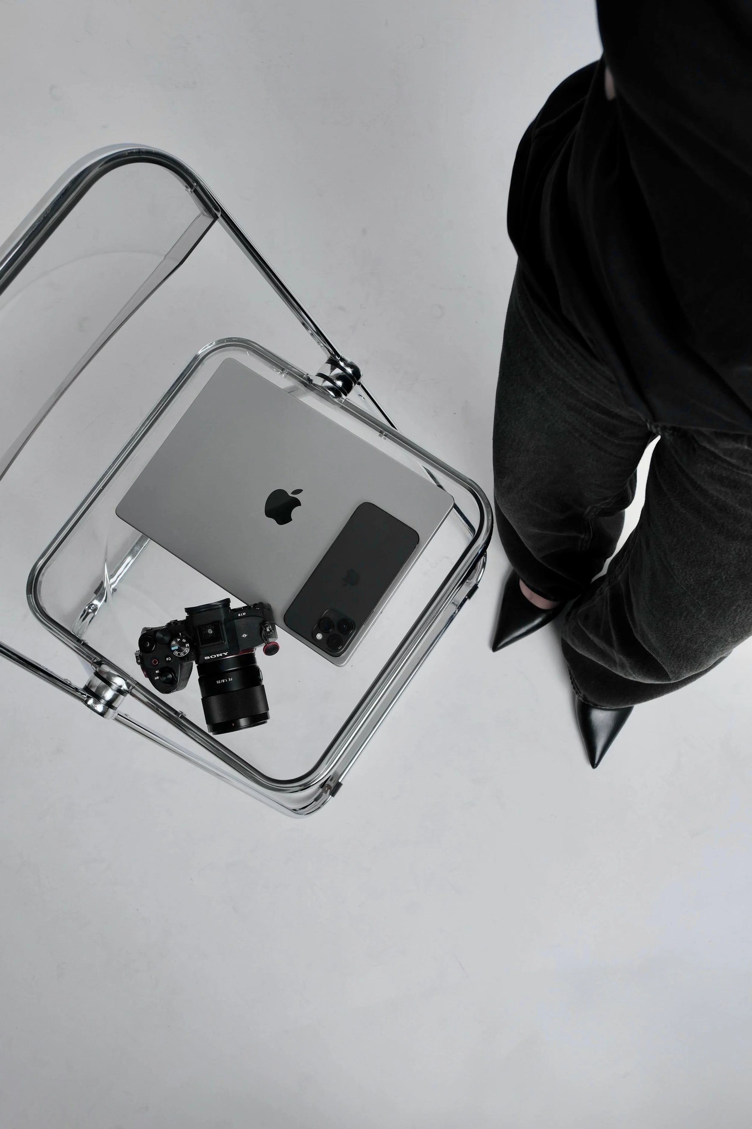 A clear acrylic chair with a glass top, placed on a white floor. On the chair, there is an Apple iPad, an iPhone, a black camera, and some black pointed shoes. A person in black pants and black shoes is partially visible, standing beside the chair.