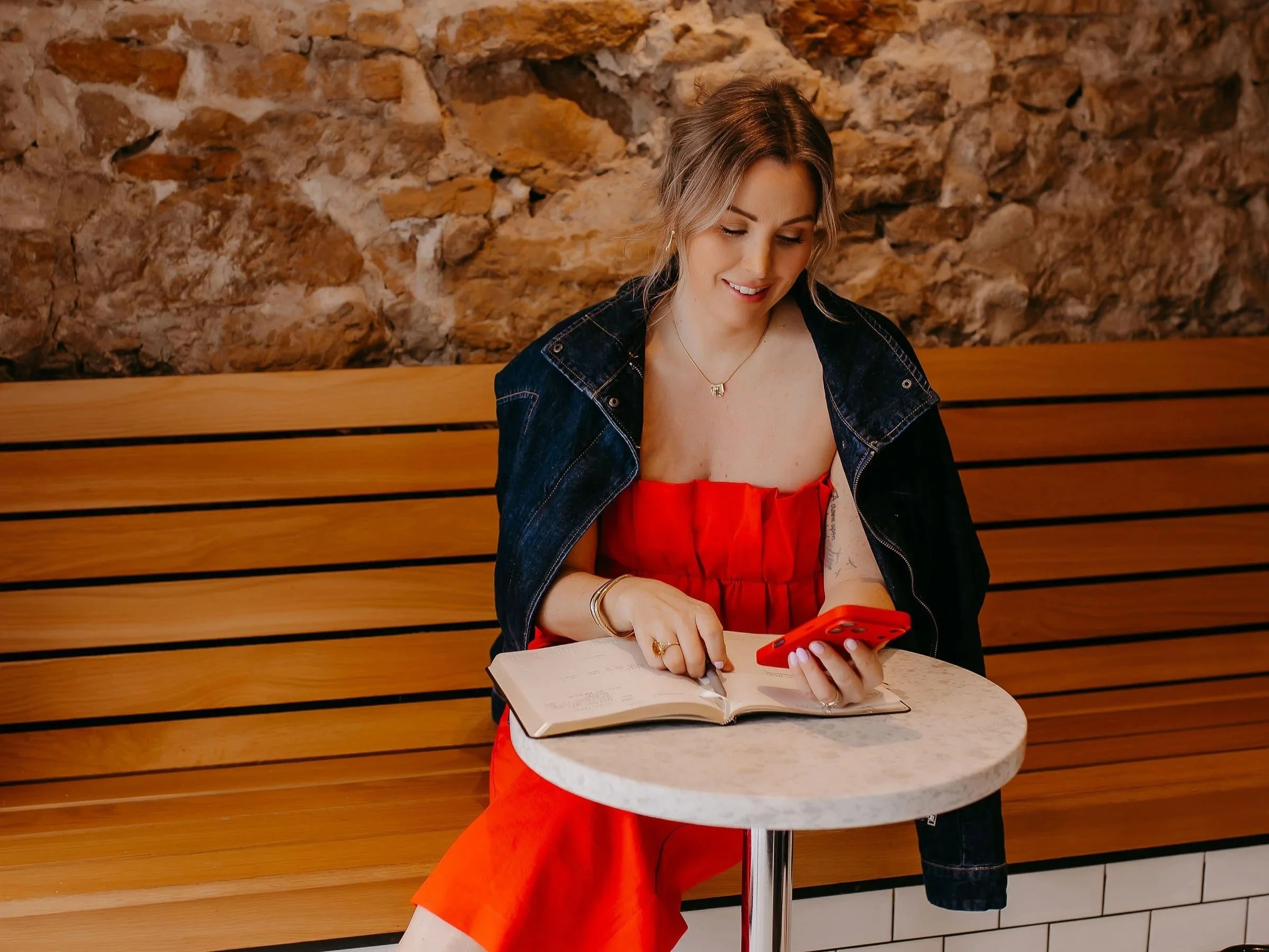 A woman in a red dress and denim jacket sitting at a small round white table, looking at her red phone, with an open notebook in front of her, against a rustic brick wall.