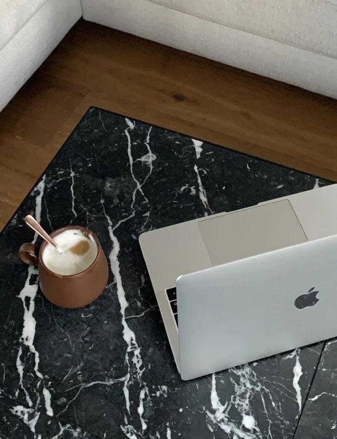 A black marble table with white veins holding a brown mug with a spoon and a frothy coffee drink, next to an open silver Apple MacBook laptop on a wooden floor near an off-white wall.