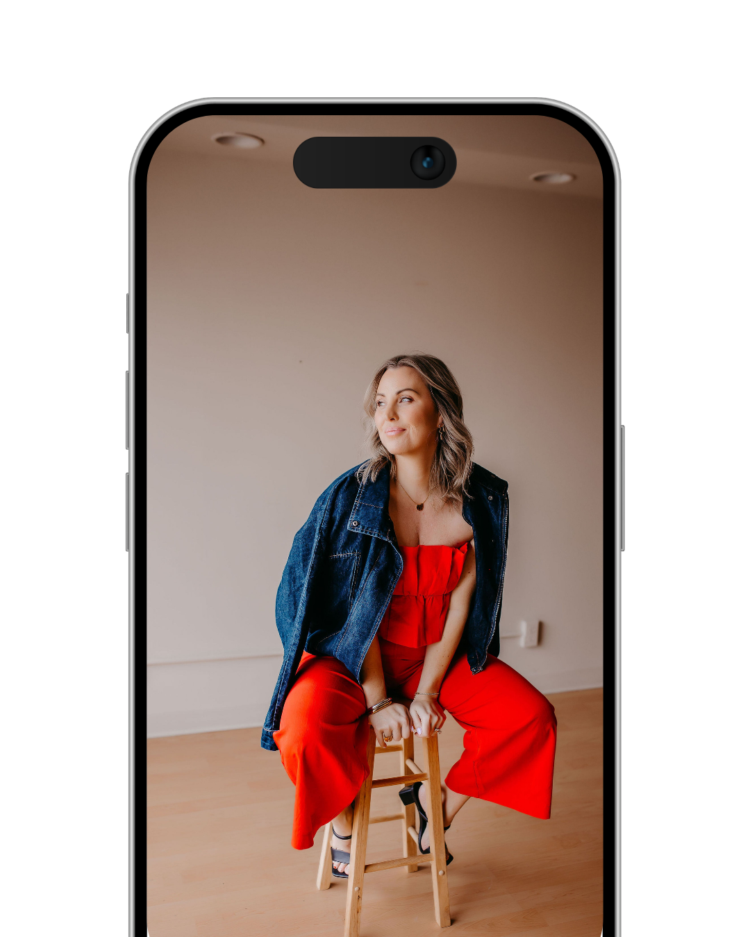 A woman sitting on a wooden stool, wearing a red outfit and a denim jacket, with a neutral indoor background.