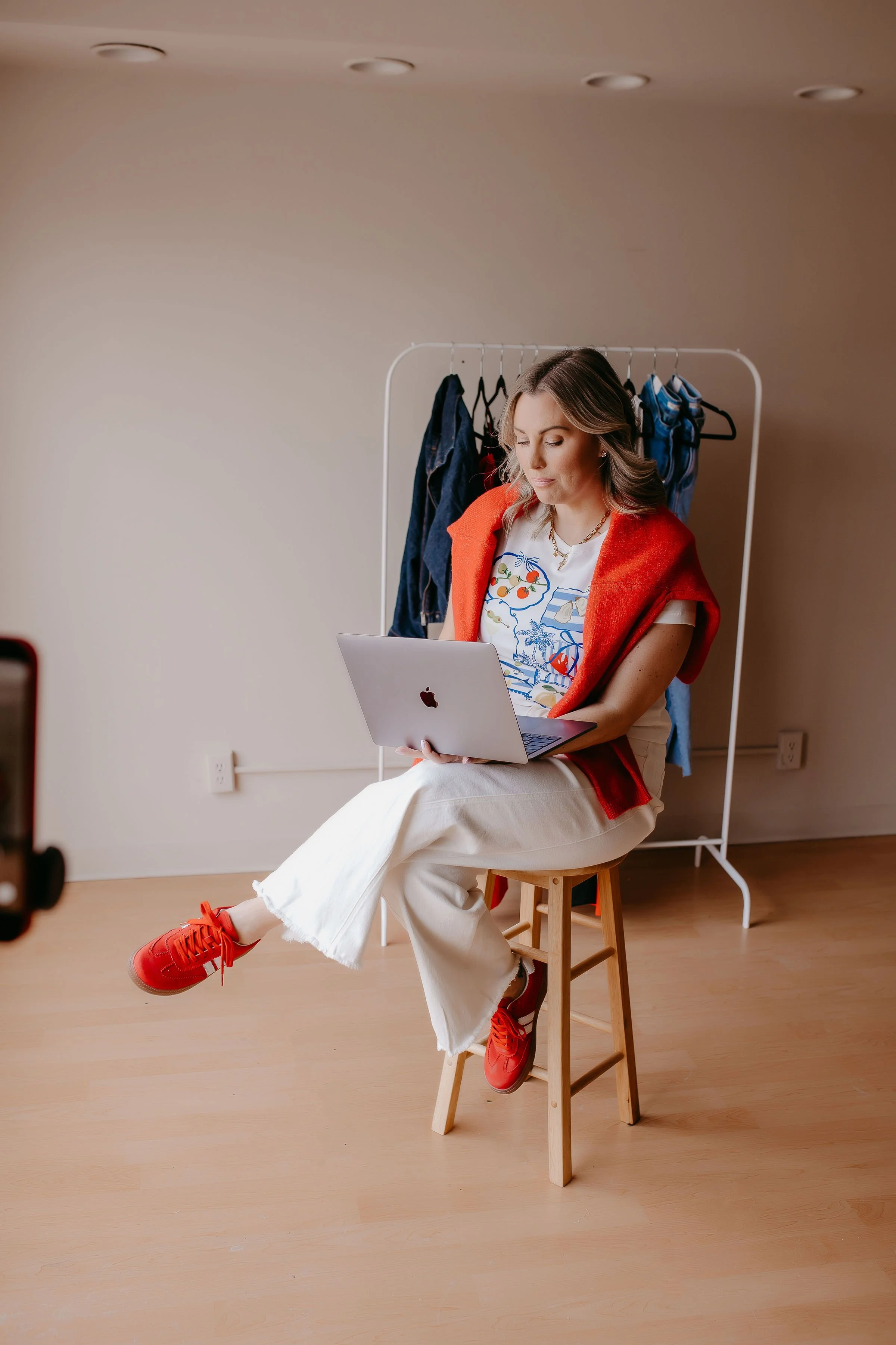 A woman sits on a wooden stool with her legs crossed, wearing red sneakers, white pants, a colorful patterned T-shirt, and a red sweater draped over her shoulders. She is working on a silver laptop in a room with a beige wall and a hardwood floor. Behind her, there is a clothing rack with jeans and other apparel hanging on it.