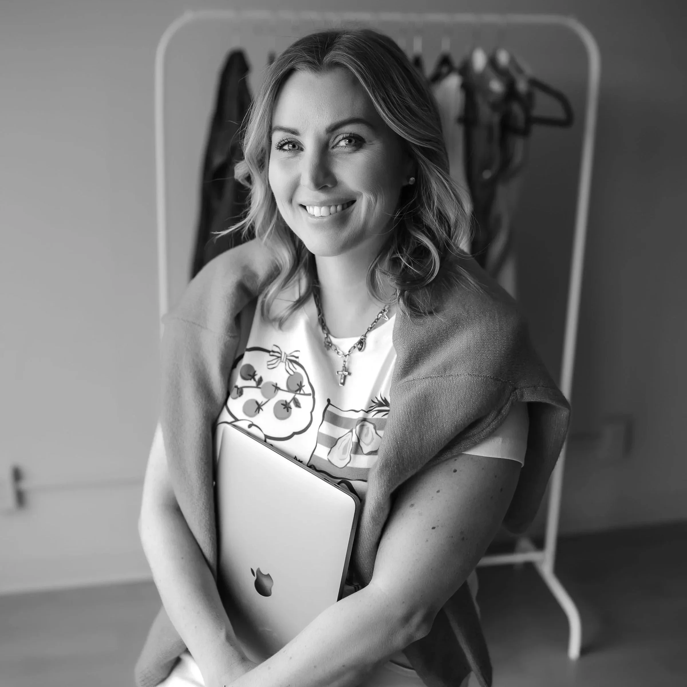 A woman with wavy hair smiling at the camera, sitting on a chair in front of a mirror with hangers visible.