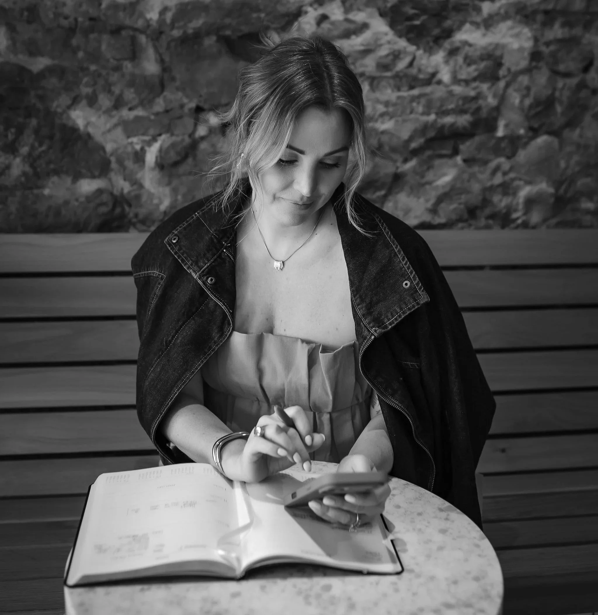 A young woman with wavy hair wearing a denim jacket, sitting at a table with an open book and a cup, looking down at her phone, with a brick wall background.