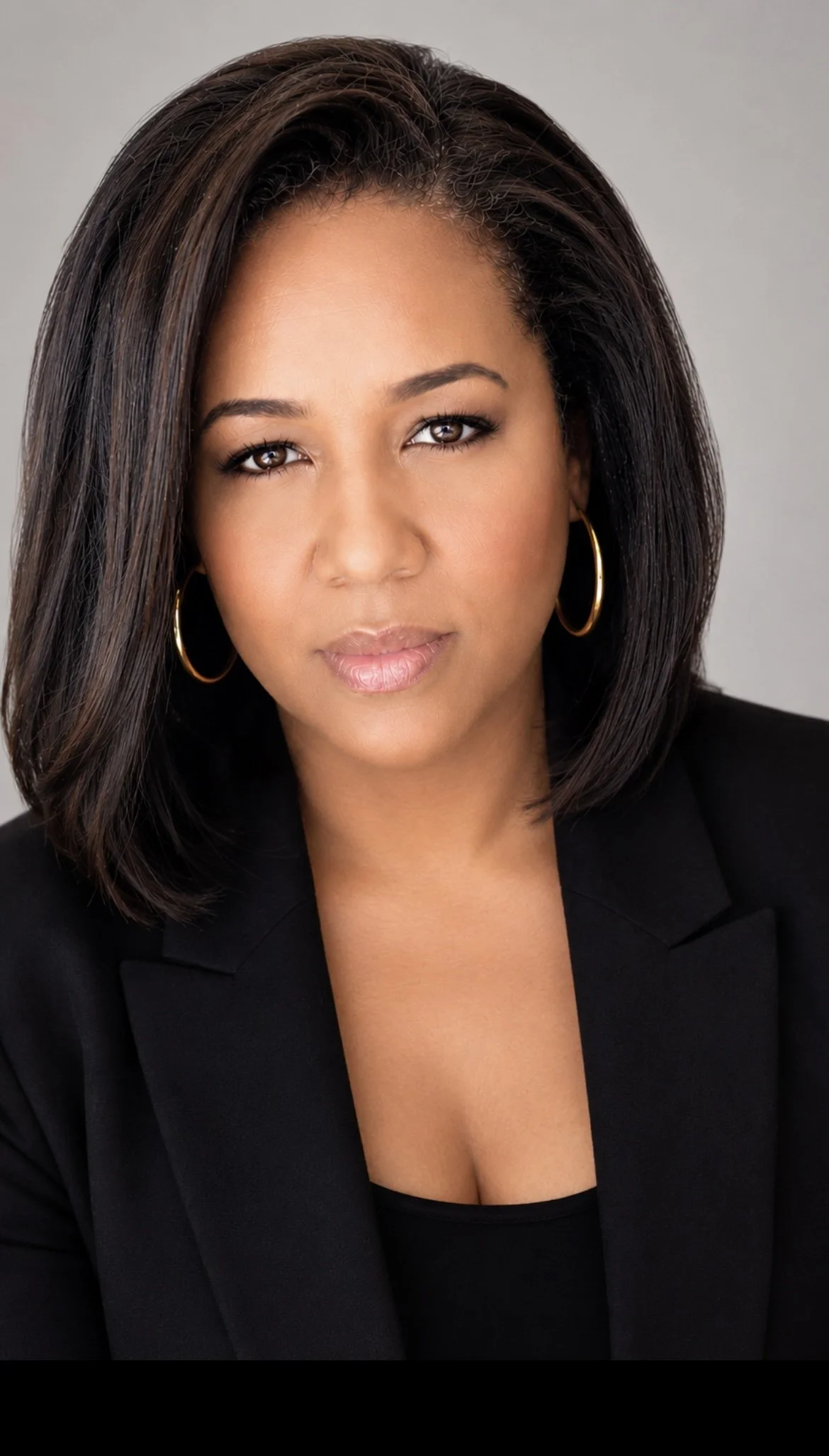 A woman with shoulder-length dark hair, brown eyes, wearing gold hoop earrings, a black blazer, and a black top, looking confidently at the camera against a plain gray background.