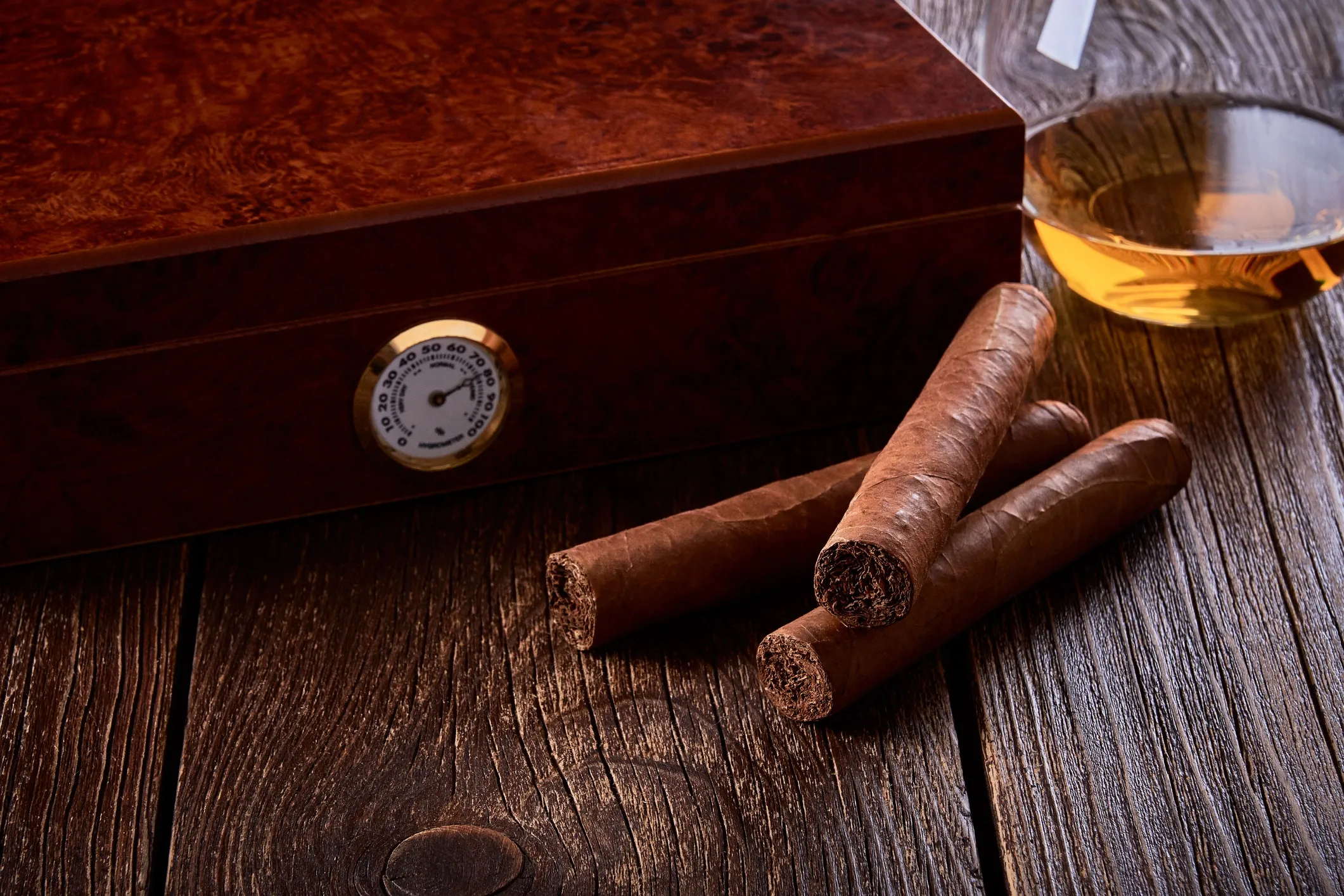 Three cigars and a glass of whiskey on a wooden surface with a wooden box in the background.