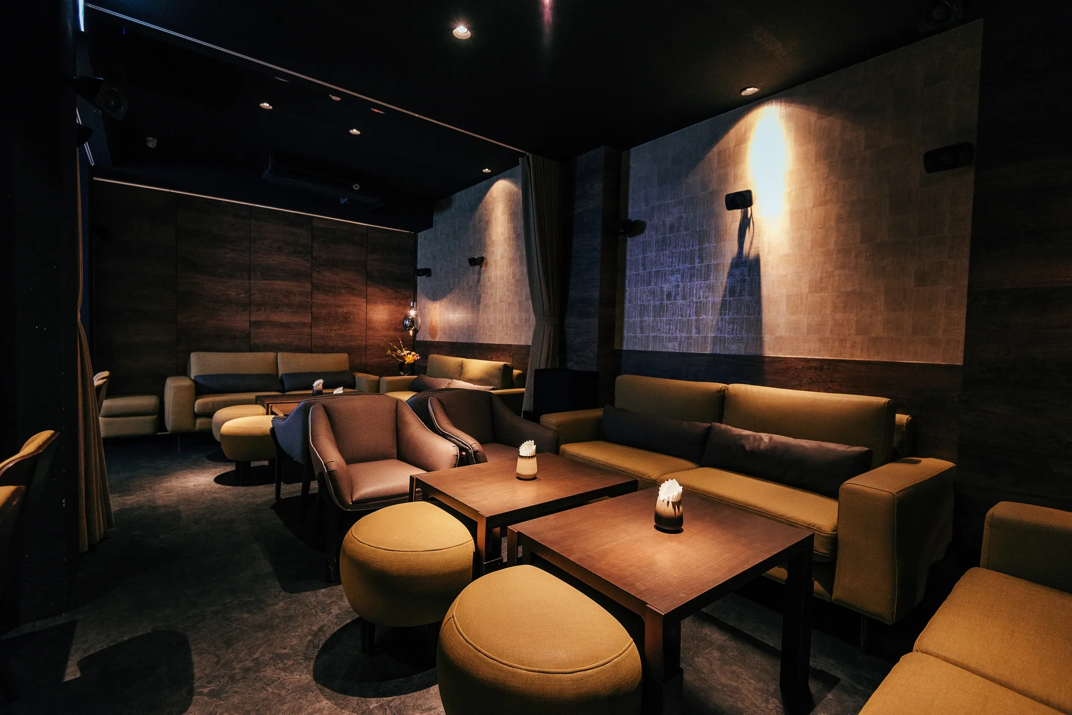 Dimly lit lounge with modern dark wood and beige seating, small wooden tables with candles, and a curtain dividing the space.