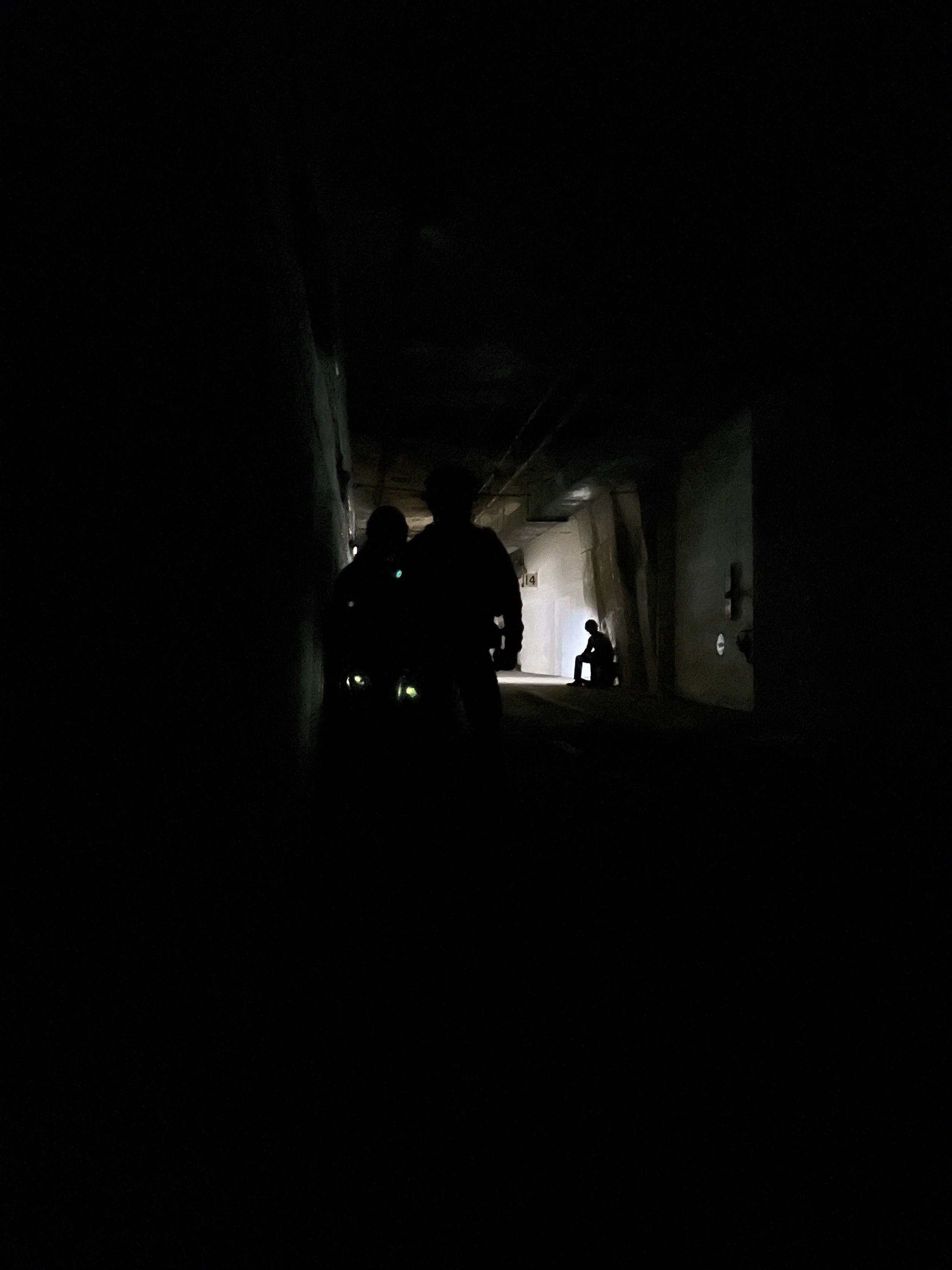 Silhouettes of two people walking through a dark, enclosed corridor with light at the far end and a person sitting against the wall