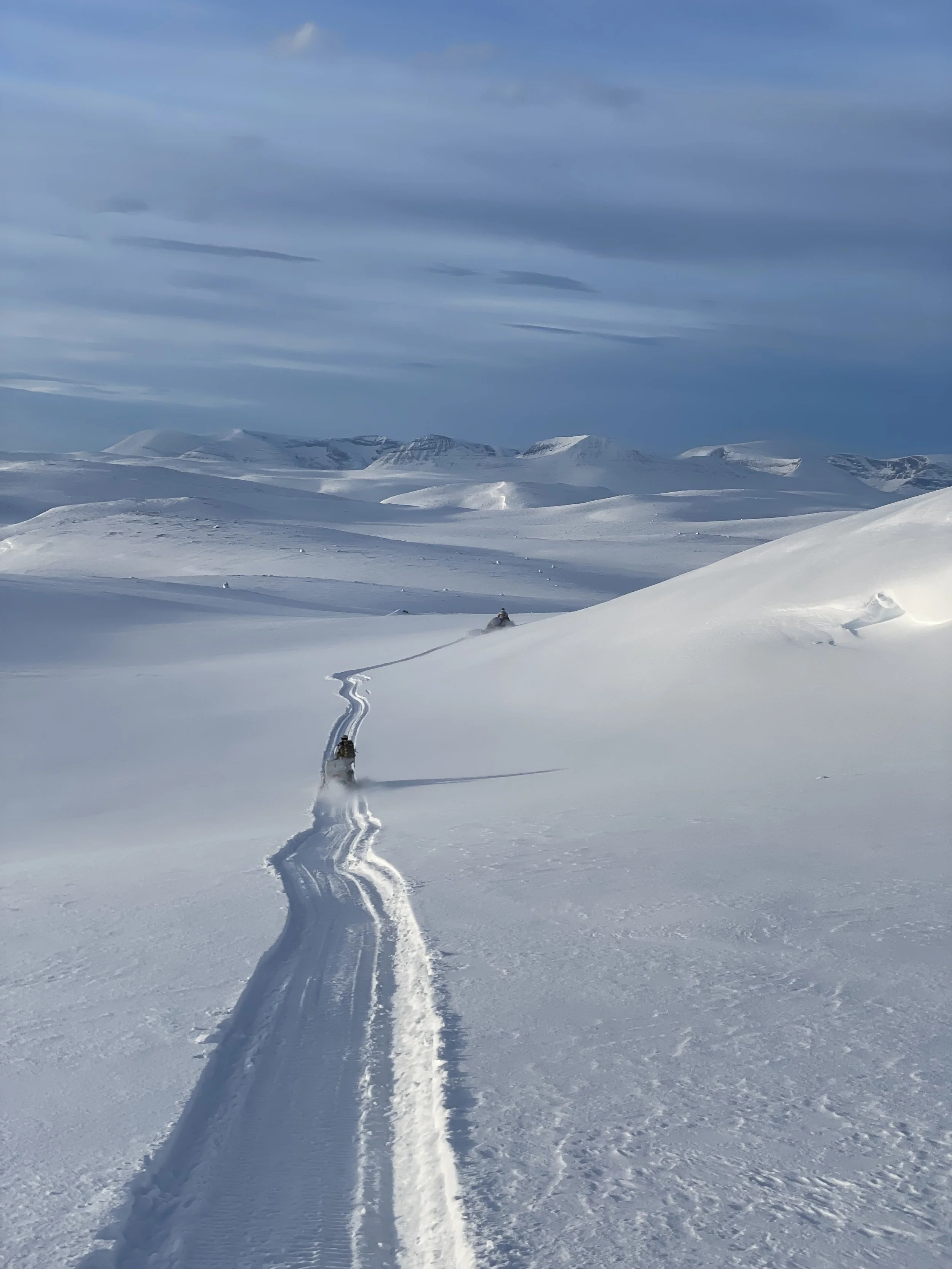 Snow-covered landscape with two snowmobiles making tracks in the snow, and distant snow-covered mountains under a cloudy sky.