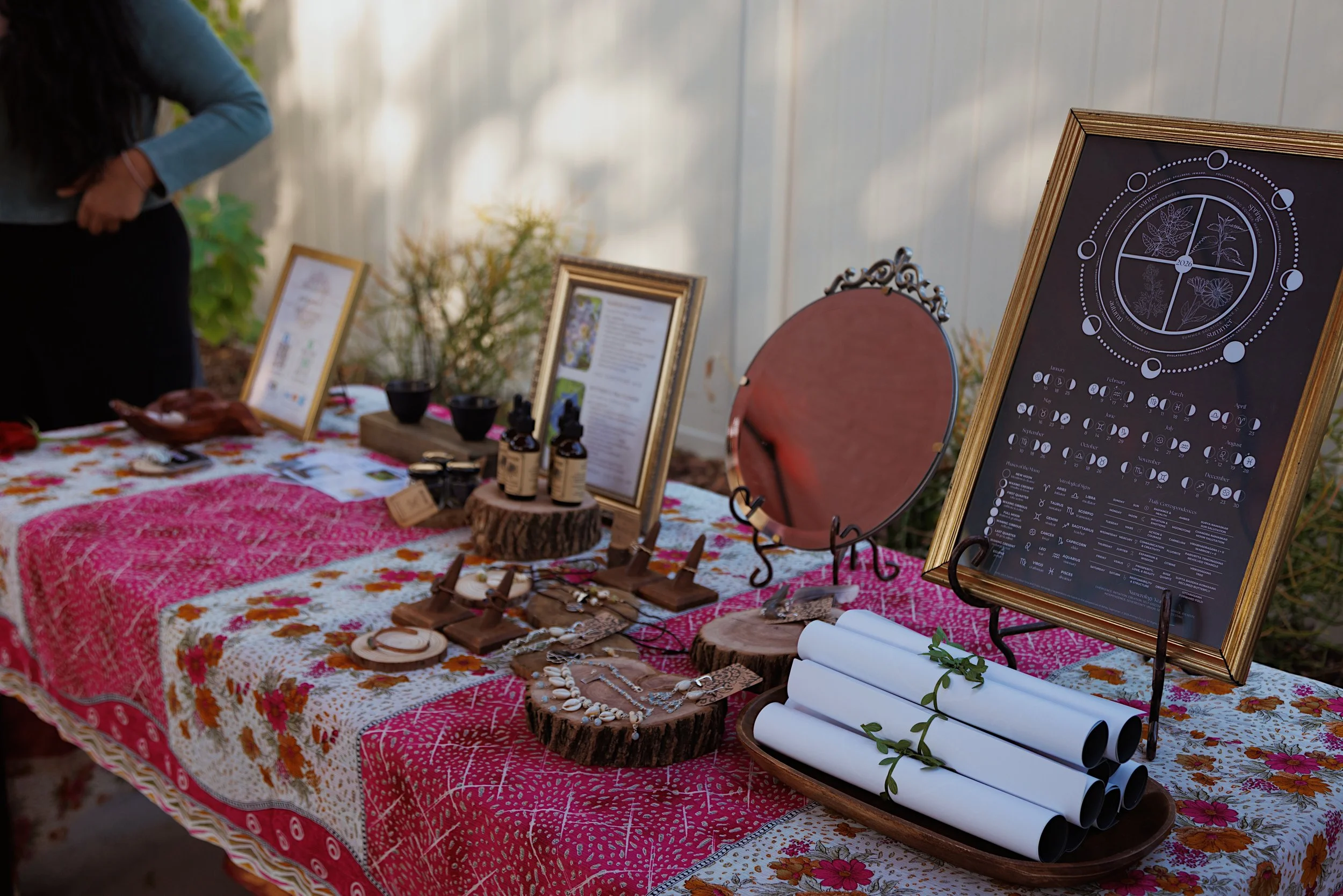 Display of herbal products, framed botanical charts, and informational boards on a table covered with a pink patterned cloth at an outdoor event.