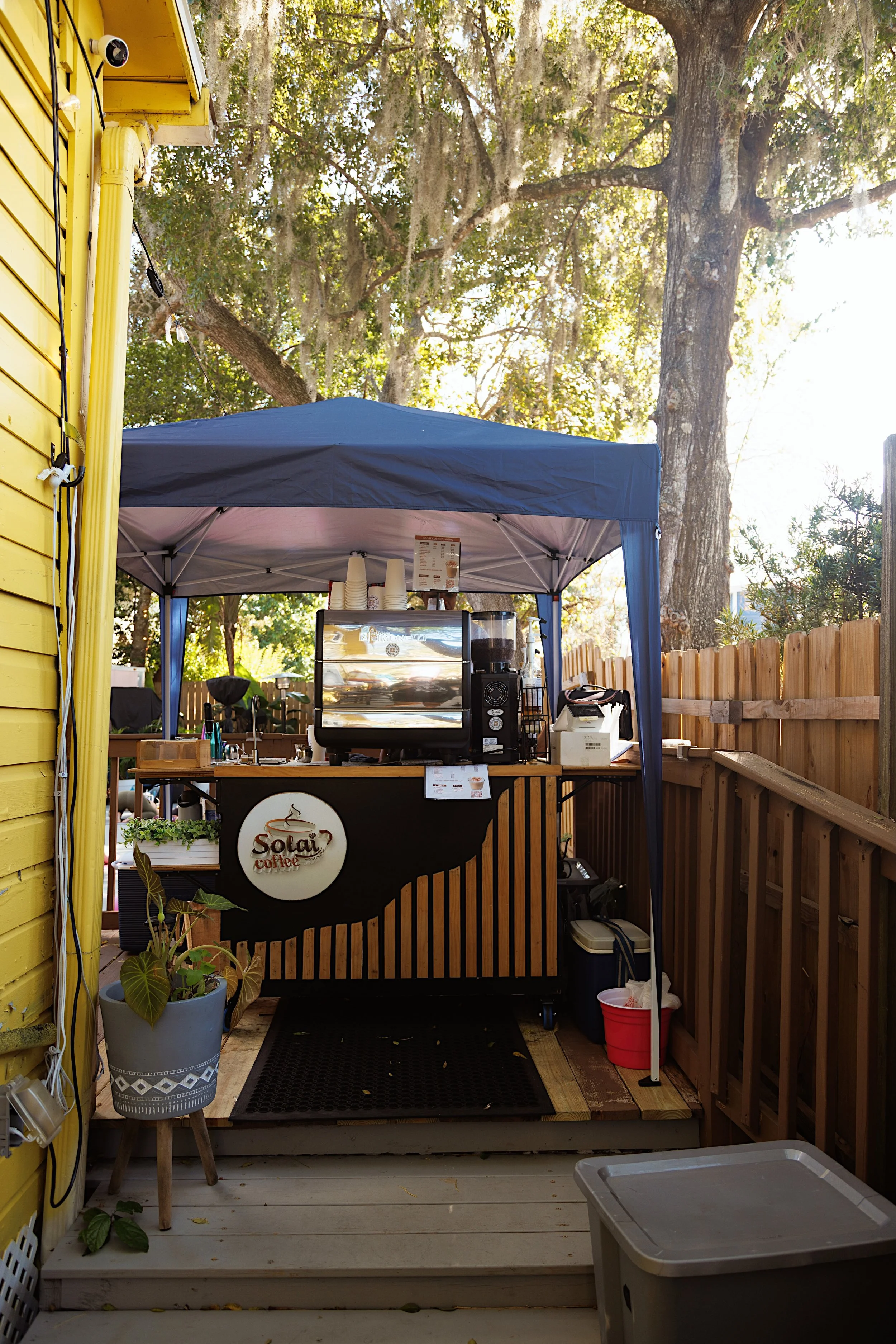 Outdoor coffee stand with a blue canopy, coffee machine, and menu, surrounded by trees and a wooden fence, with plants on the porch and outdoor storage items.