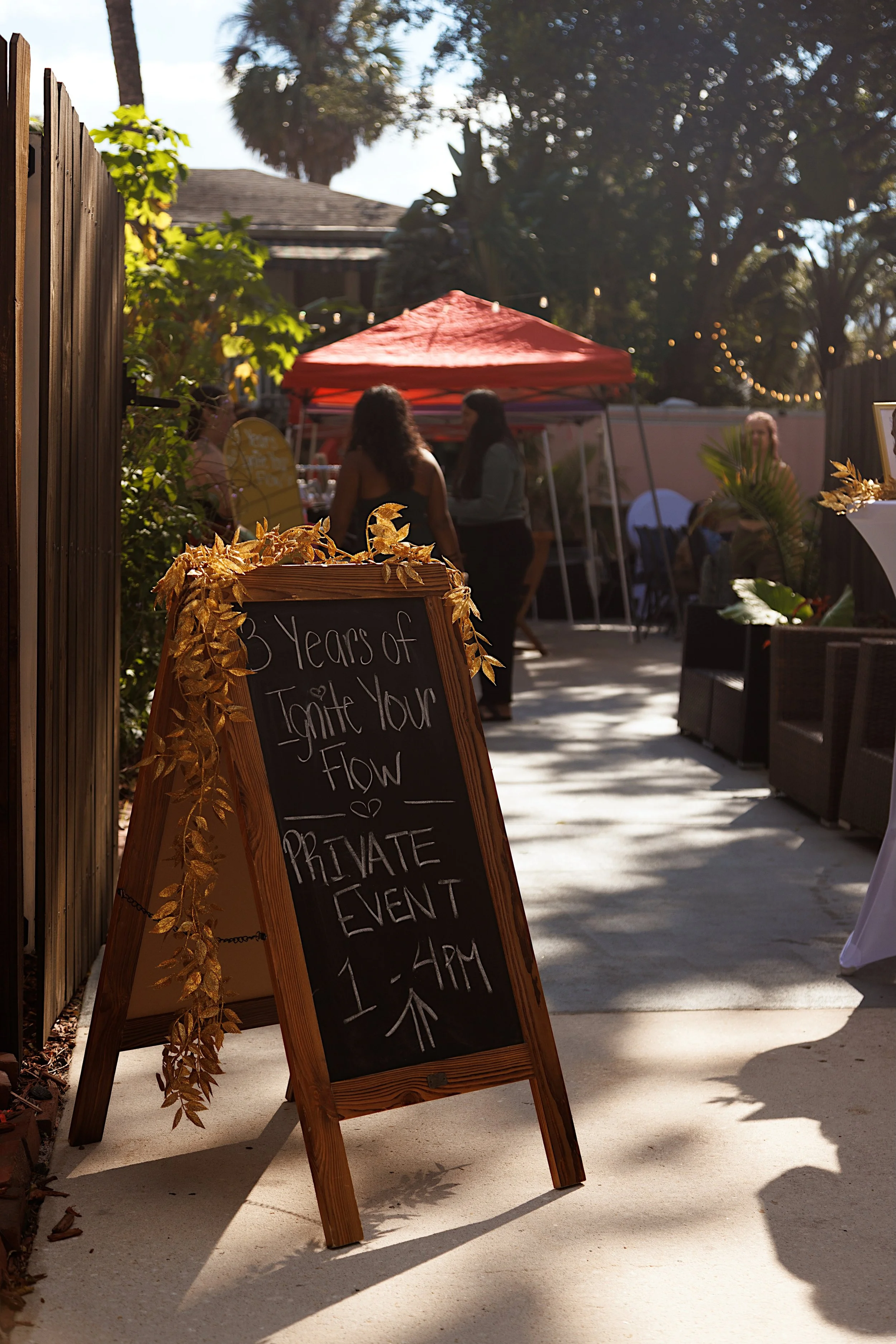Outdoor private event sign with gold leaves decoration, advertising an event celebrating 3 years of something, with the time from 1 to 4 PM. In the background, there are people socializing under a red canopy, trees, string lights, and a wooden fence.