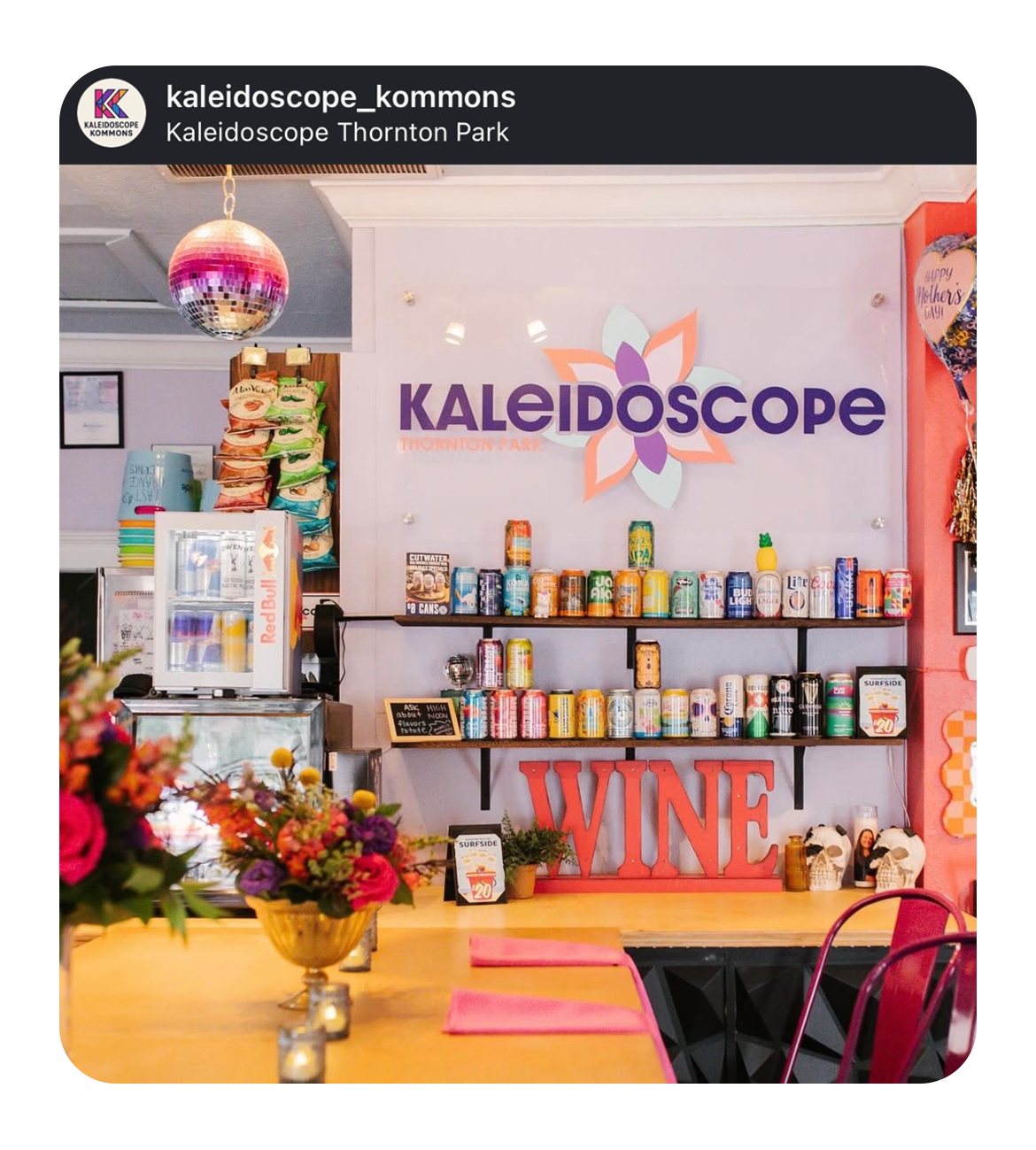 Interior of Kaleidoscope Thornton Park with a wall decorated with a large sign that says 'Kaleidoscope' with a flower motif, shelves filled with beverages, and a table with a flower arrangement in the foreground.