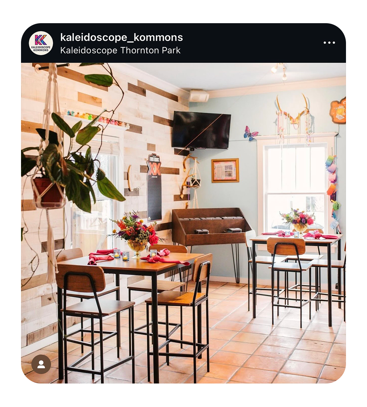 Interior of a decorated dining area at Kaleidoscope Thornton Park with wooden tables, chairs, and floral centerpieces, featuring a pinball machine and a window with pink curtains in the background.