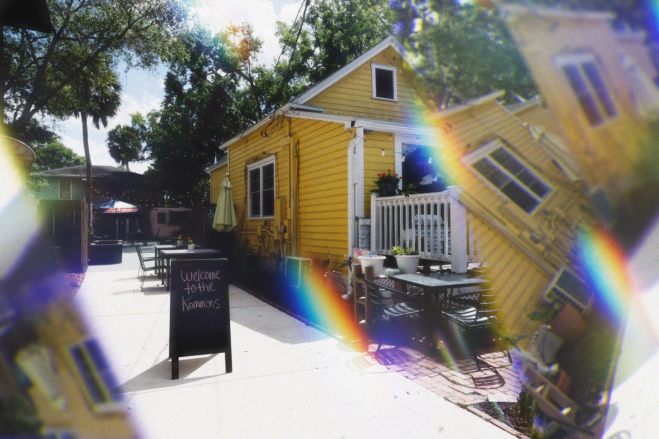 Kaleidoscope Thornton Park, a yellow building with white trim and a small porch, outdoor patio with tables and chairs, potted plants, sign that says 'Welcome to the Kommons', trees and partly cloudy sky in the background.