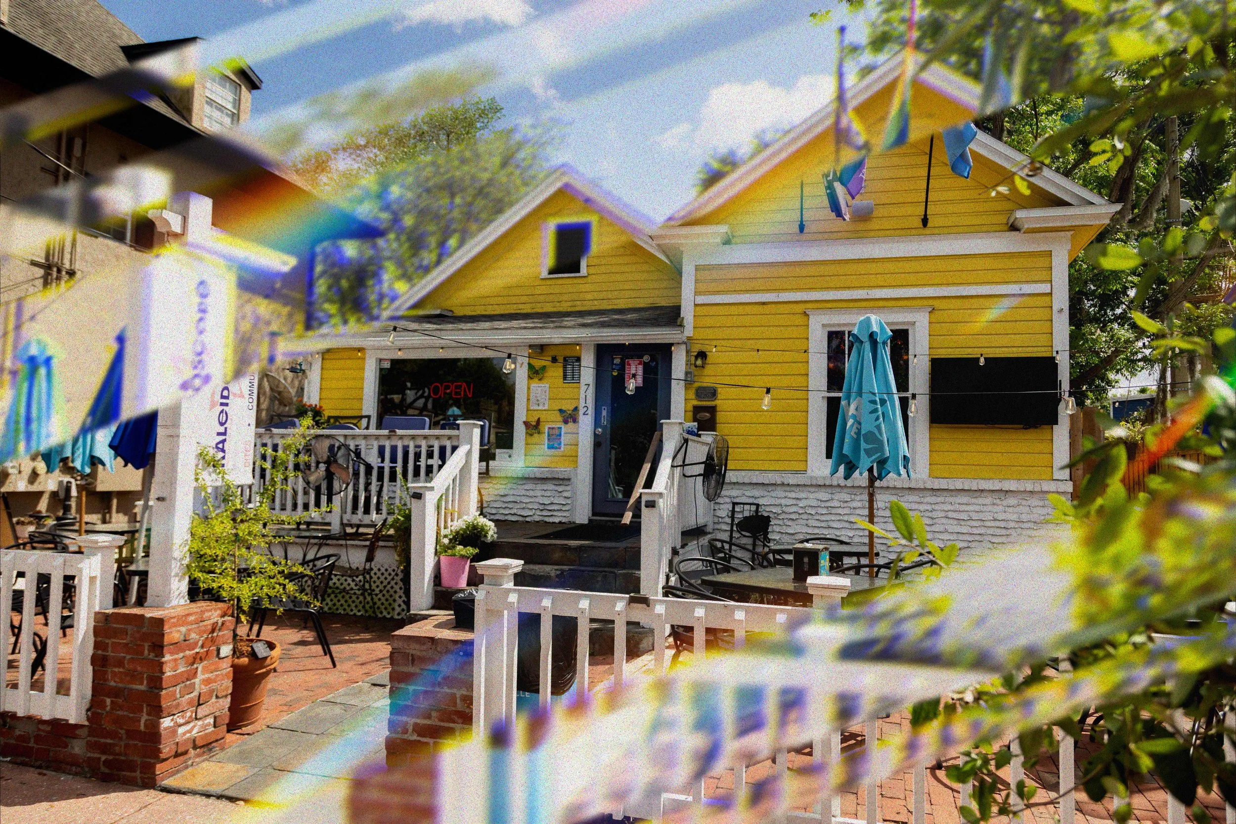 Front, street view of Kaleidoscope Thornton Park, which is yellow house with a front porch, part of an outdoor seating area with tables, chairs, and umbrellas, and string lights. .