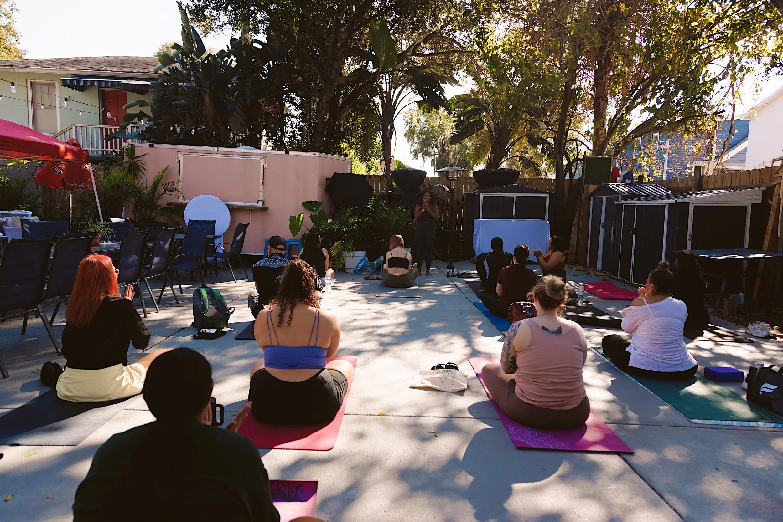 Group of people participating in outdoor yoga in the back Kaleidoscope Thornton Park, called The Kommons, with trees, a fence, and a small building in the background.