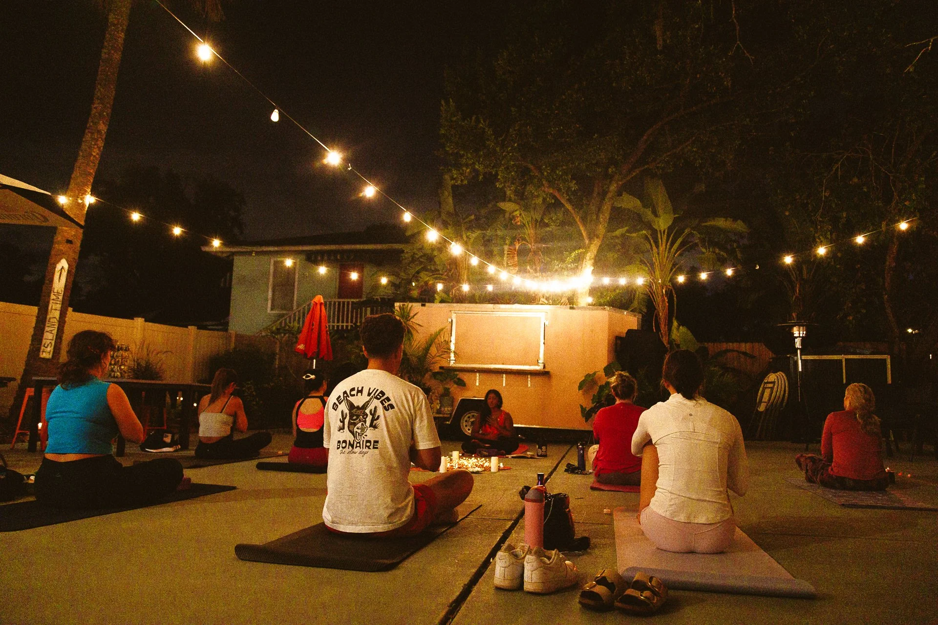 People practicing yoga or meditation during an outdoor evening gathering, sitting on mats around candles, with string lights and trees illuminating the scene.
