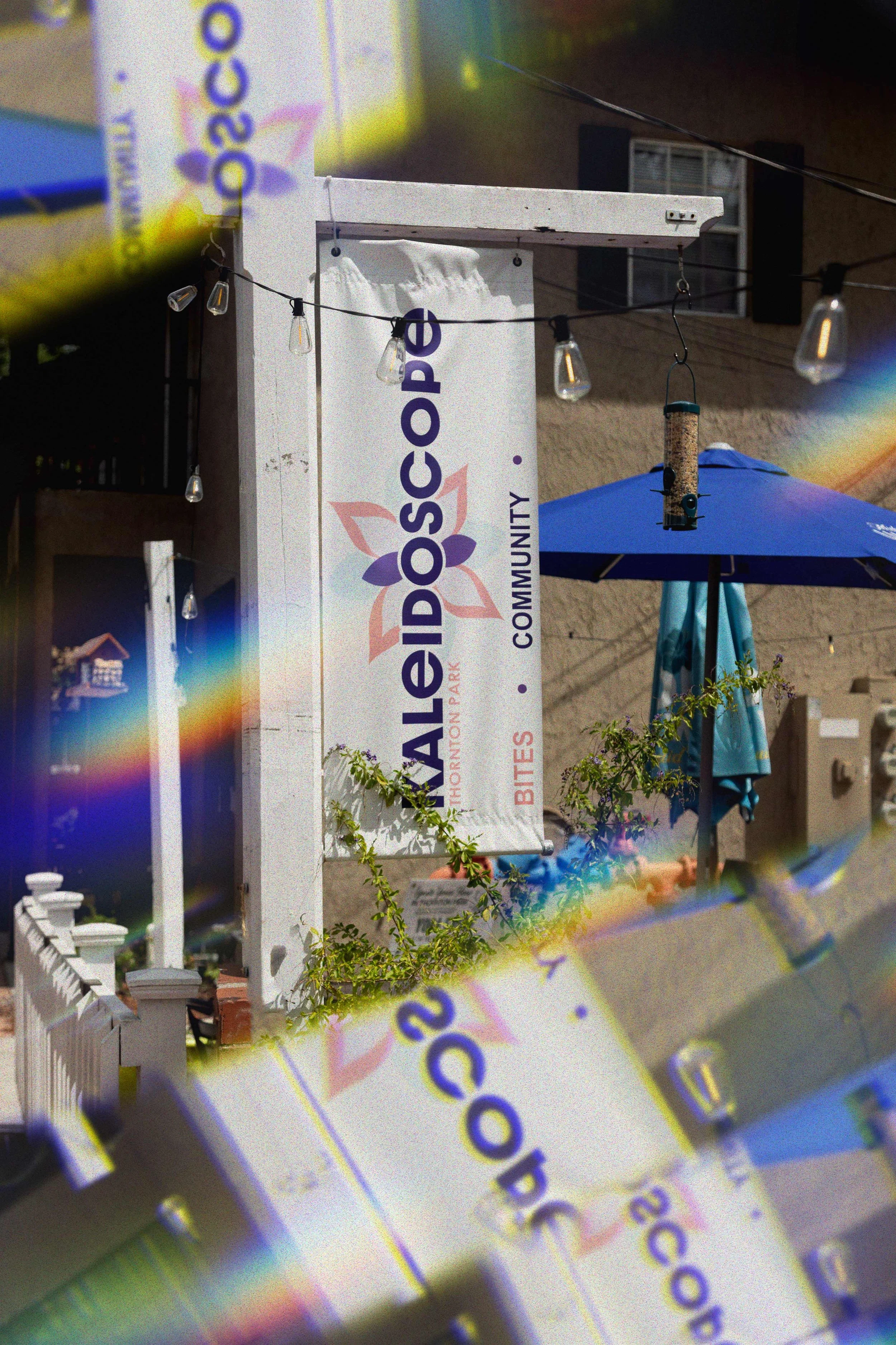 A sign for Kaleidoscope in Thornton Park in Orlando, Florida with purple and orange design, hanging on a white post outside a building, next to a blue patio umbrella, with string lights and bird feeder hanging above.
