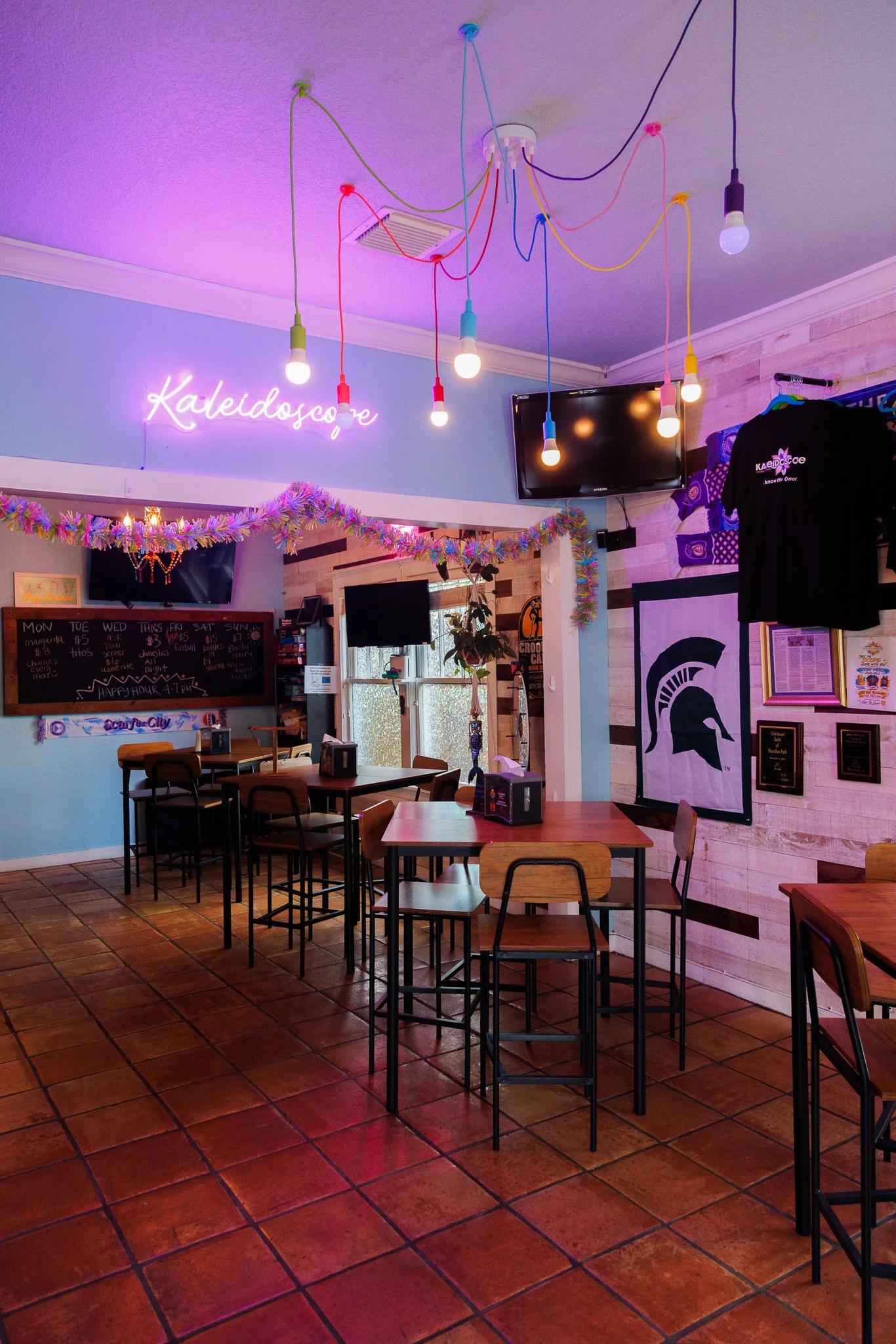 Interior view of Kaleidoscope Thornton Park with wooden tables and chairs, decorated with a purple neon sign that reads 'Kaleidoscope', colorful string lights on the ceiling, a flat-screen TV, and framed artwork on the wall.