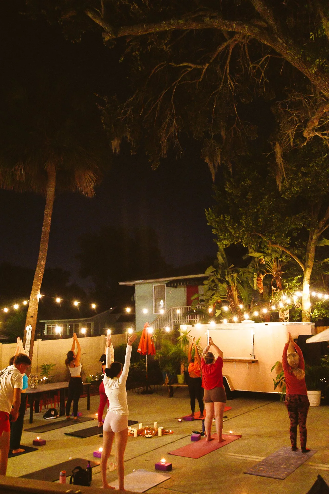 People practicing yoga outdoors at night, illuminated by string lights, with trees and a house in the background.