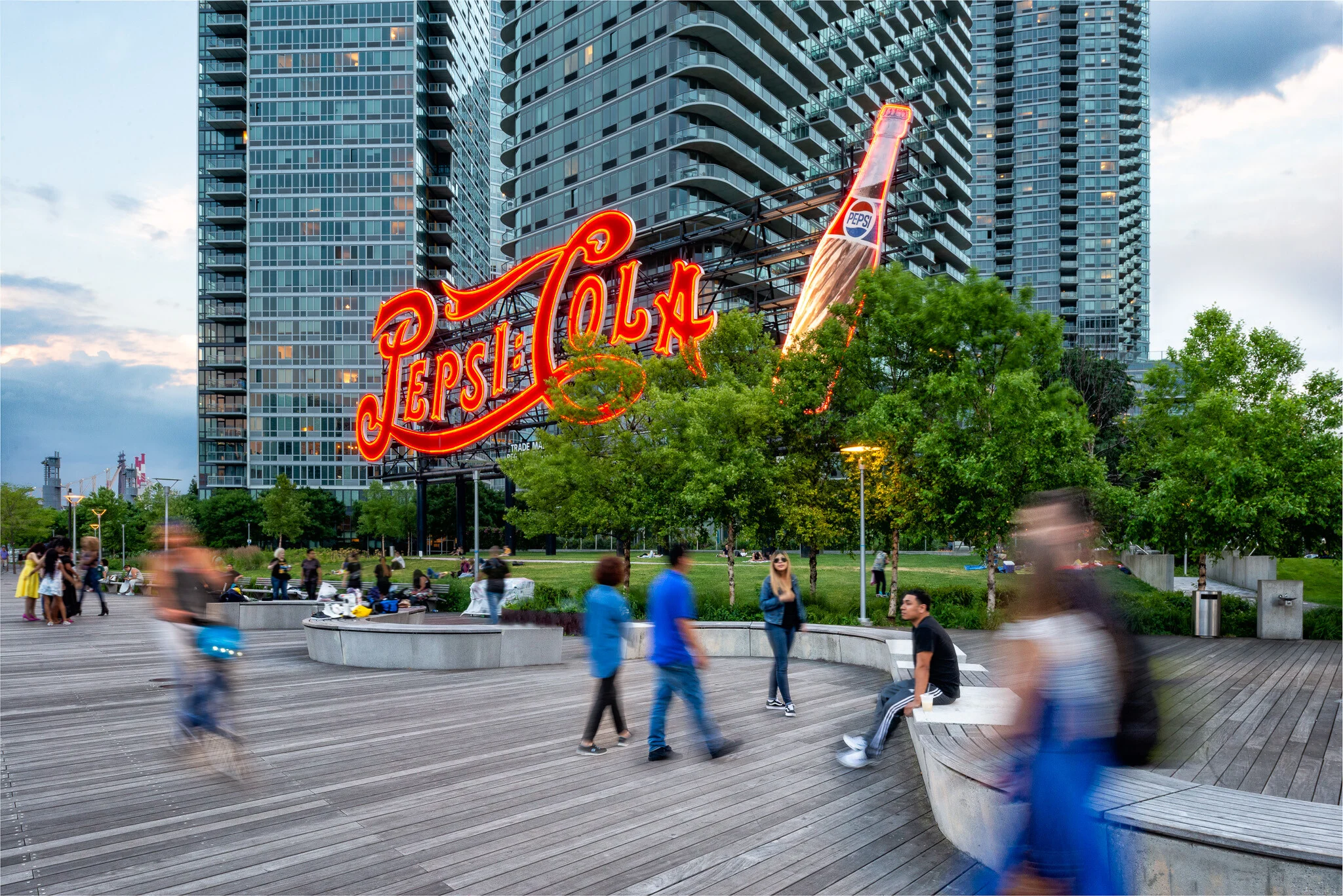 Outdoor urban park with modern high-rise buildings, trees, wooden walkways, and a neon Pepsi sign during early evening; people walking, sitting, and socializing.