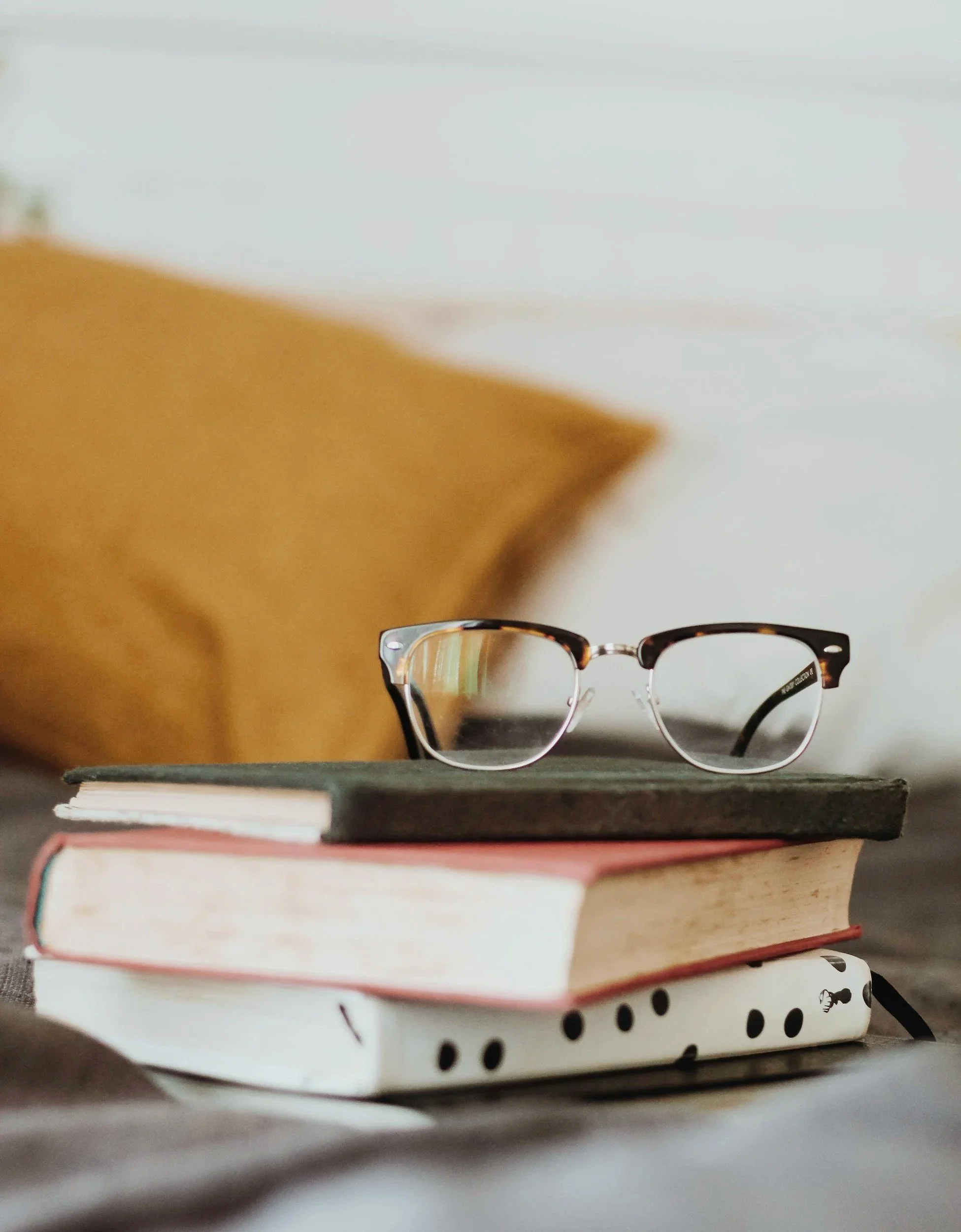 A stack of  three colorful books sits on the edge of a bed with a pair of glasses sitting on top.
