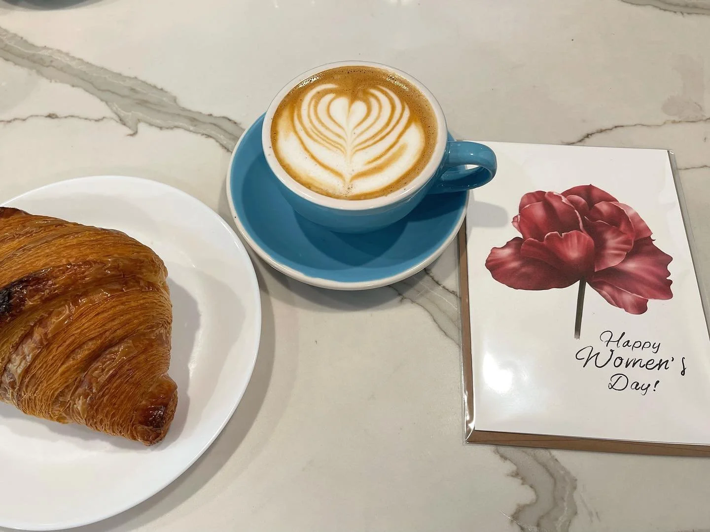 A croissant on a white plate, a latte with latte art in a blue cup and saucer, and a greeting card with a red flower and the text "Happy Women's Day!" on a marble table.