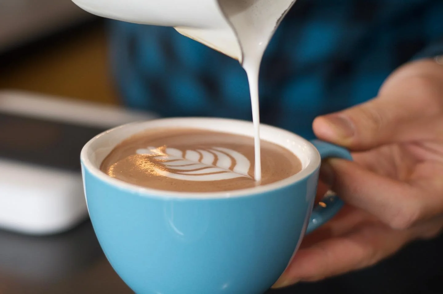 A barista pours steamed milk into a cup of coffee, creating latte art with a symmetrical pattern.