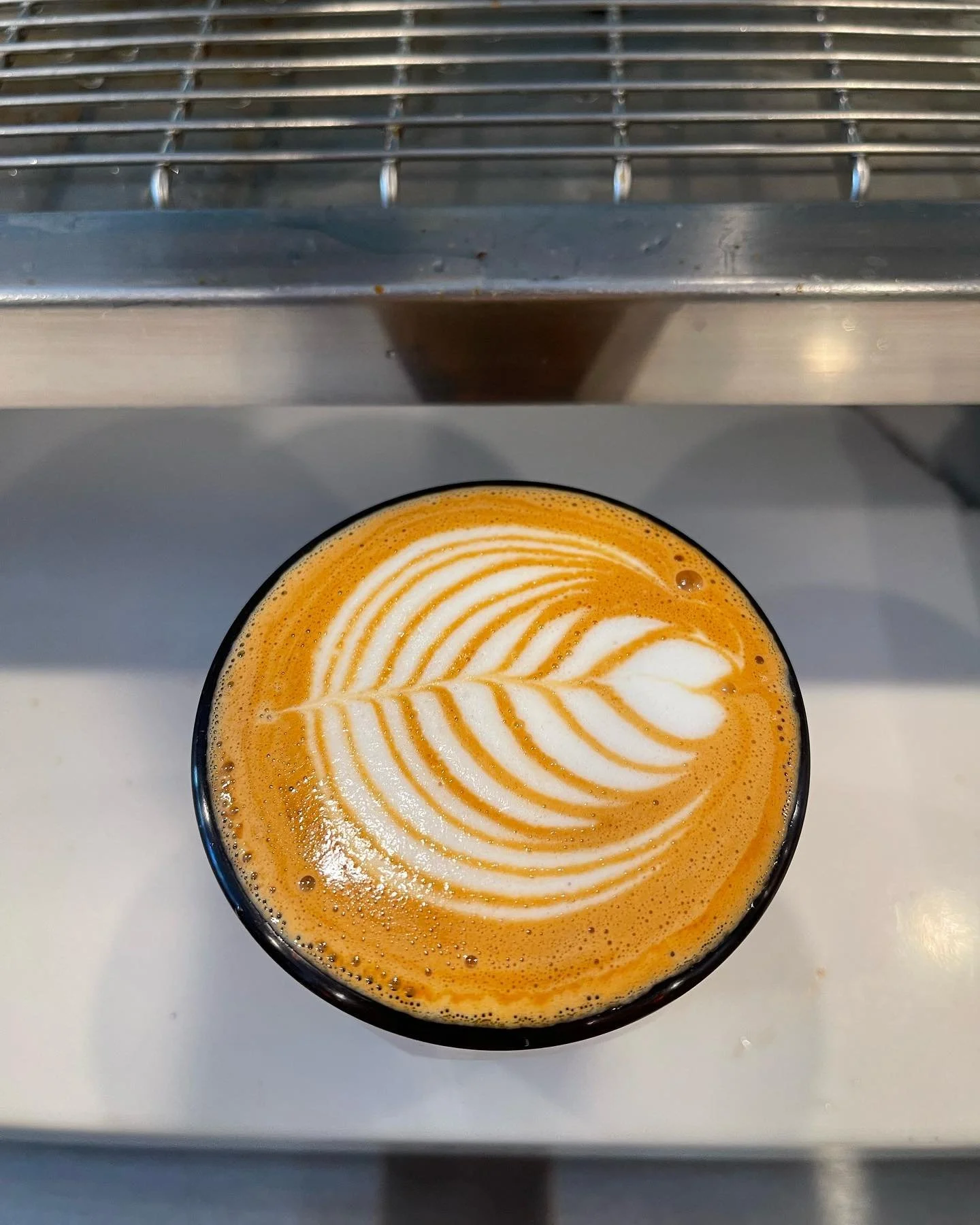A cup of coffee with latte art leaf design on top, sitting on a white surface with a metal shelf above.