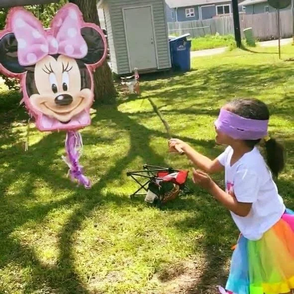 A young girl in a rainbow skirt and purple blindfold is playing with a Minnie Mouse balloon on a grassy yard. The girl is holding the balloon's string, and there are shadows of trees on the ground. A small dog and a blue trash bin are in the background.